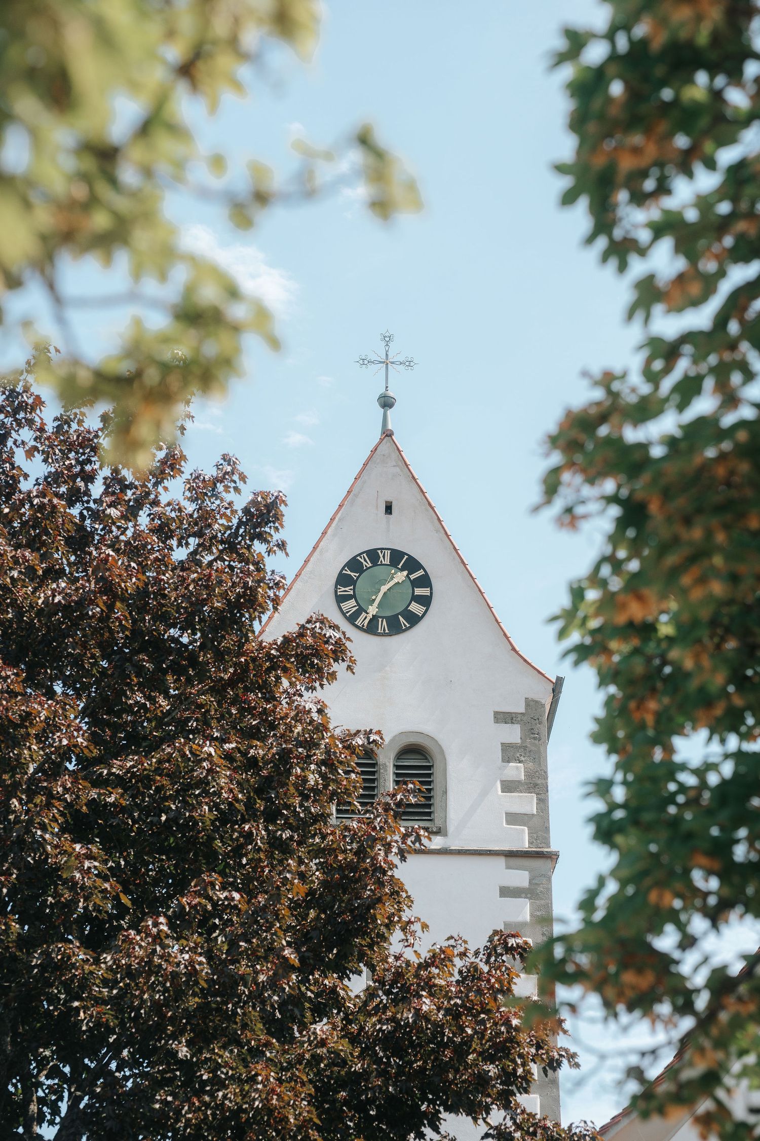 White church steeple with a green clock face and a cross on top, framed by autumn tree leaves against a soft blue sky.