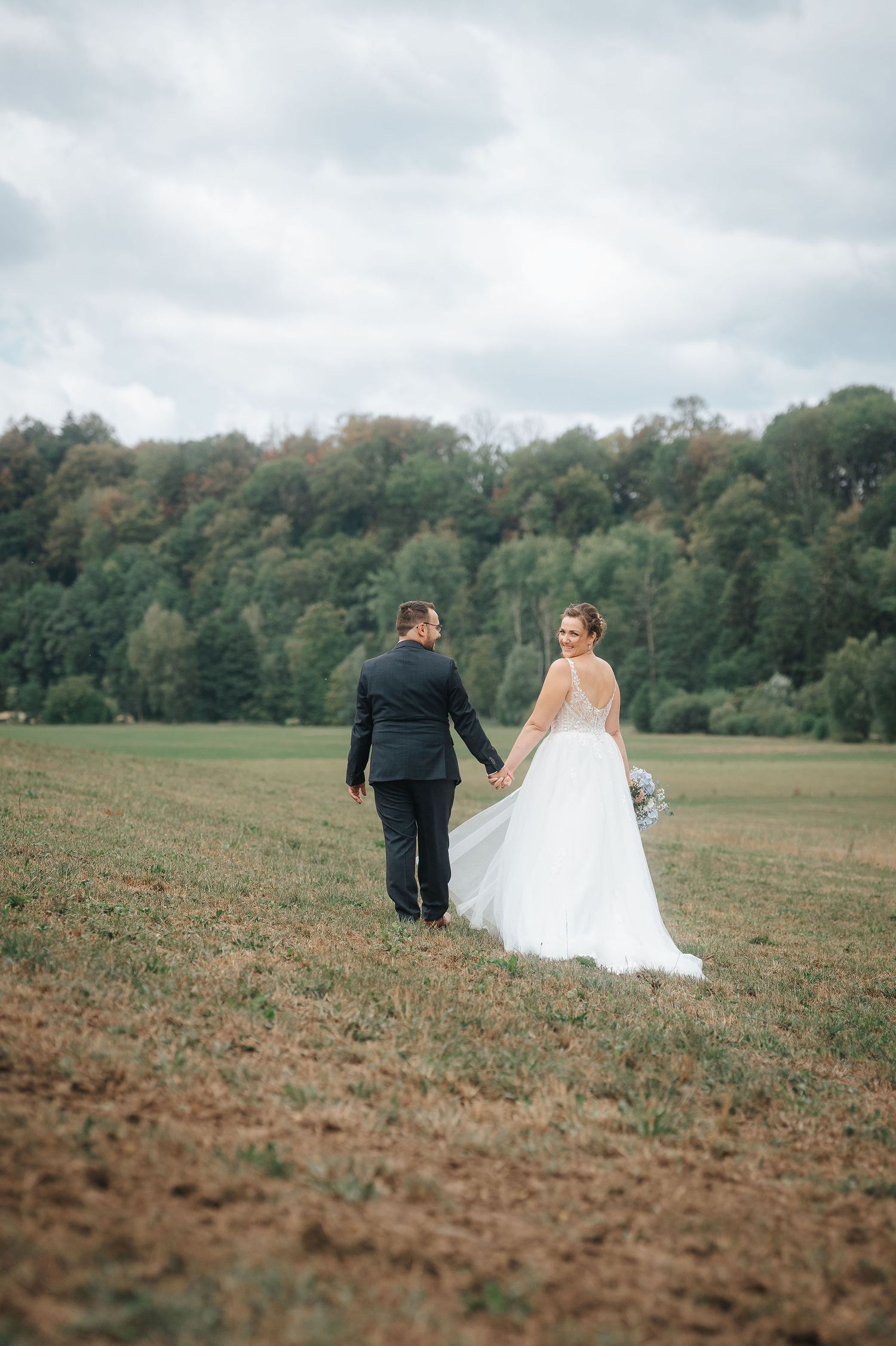 Bride in a white gown and groom in a dark suit hold hands while walking through a grassy field toward a tree-lined forest on a cloudy day.
