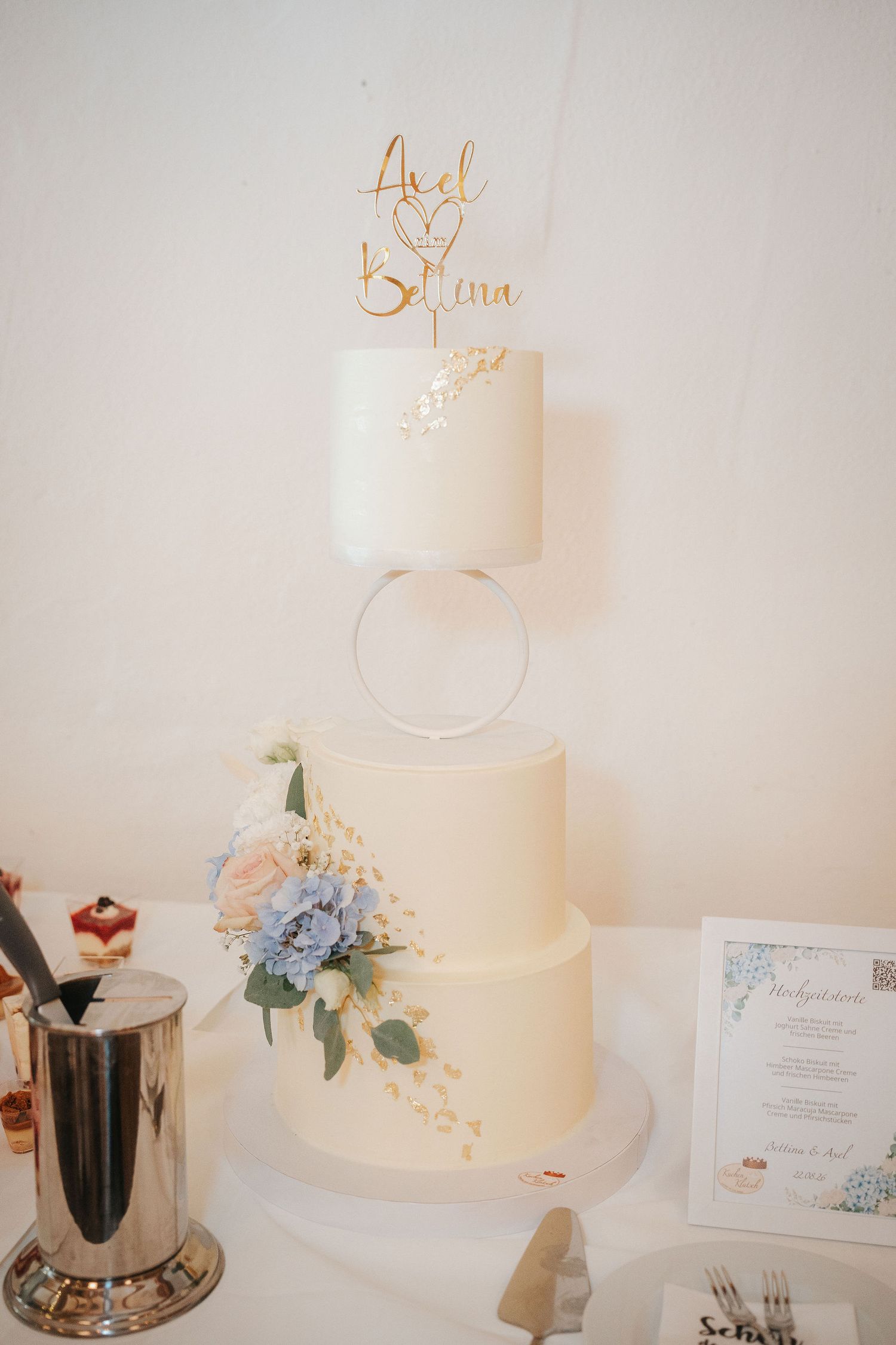 Three-tier white wedding cake with blue and blush flowers, a floating ring tier, and a gold name topper, displayed beside a menu card.