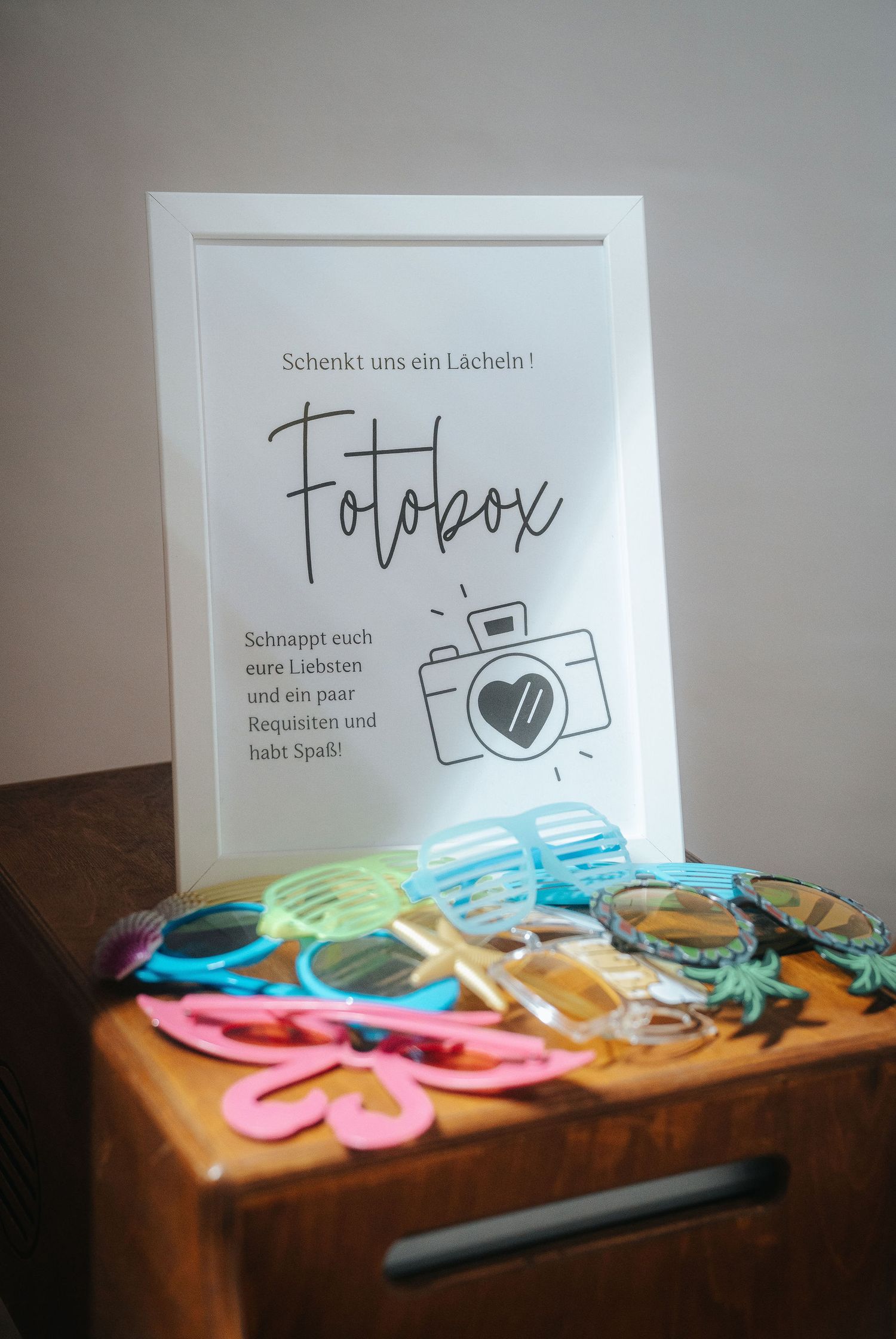 Framed German Fotobox sign with camera icon sits on a wooden cabinet beside colorful photo booth props, sunglasses and party glasses.
