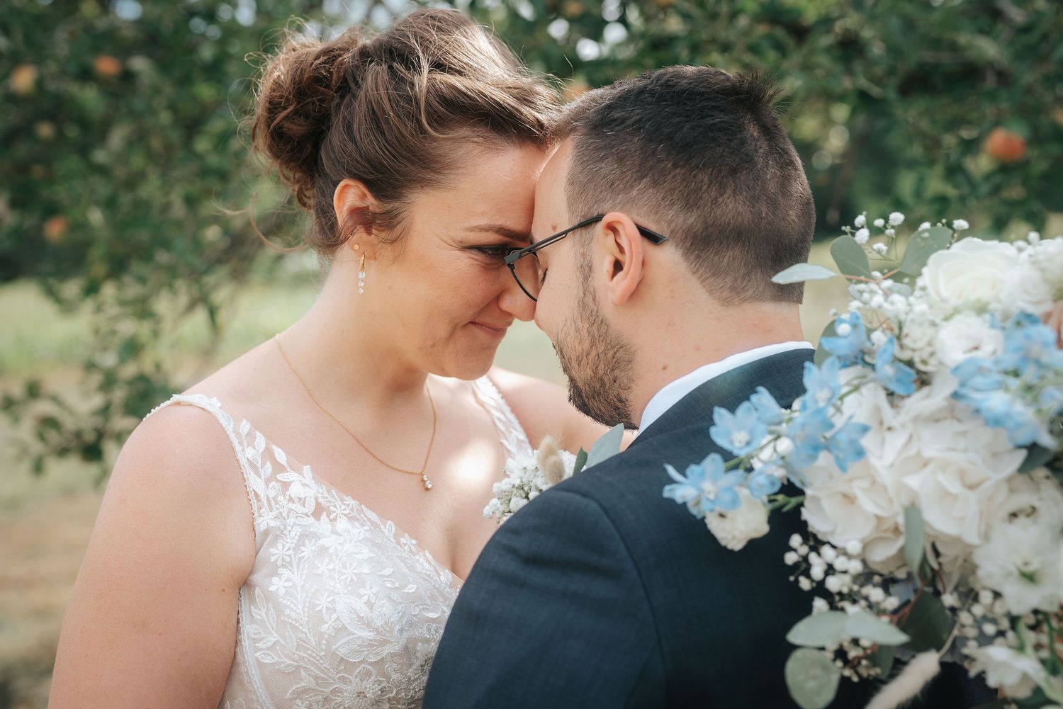 Bride and groom touch foreheads in a tender moment outdoors, holding a bouquet of white and blue flowers in an orchard.