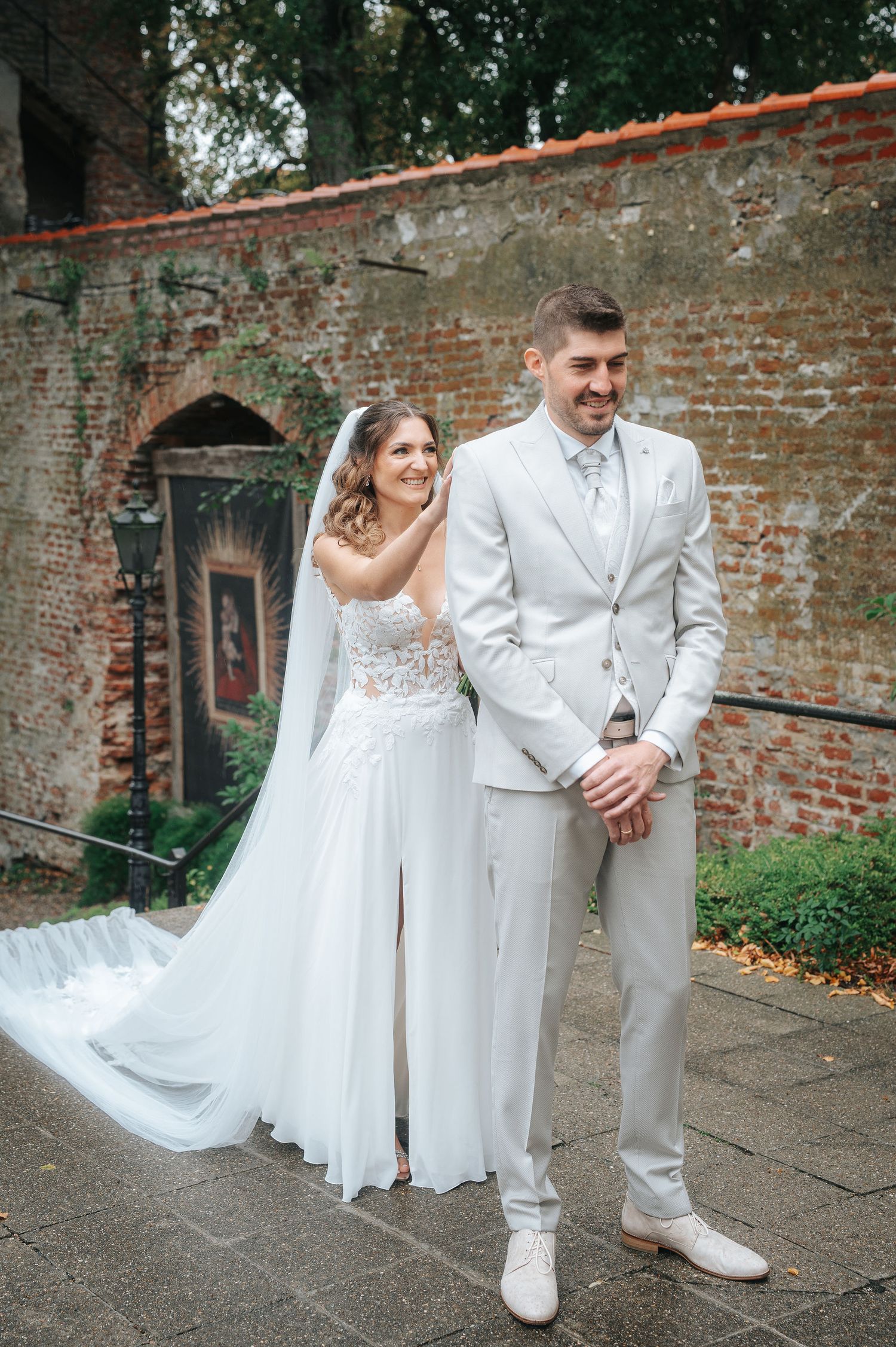 Bride in a lace wedding gown and long veil touches the shoulder of a smiling groom in a light gray suit beside an old brick wall.
