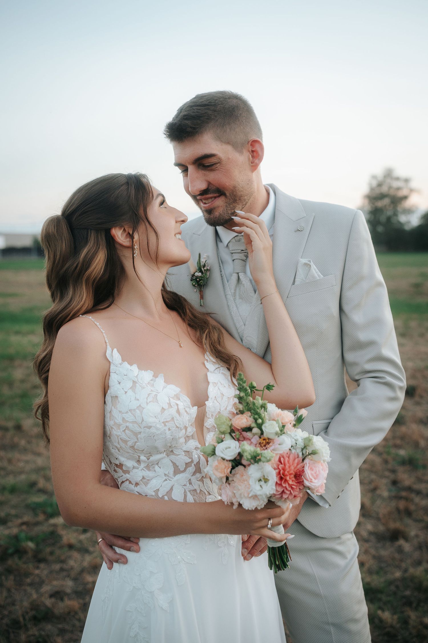 Smiling bride in a white lace gown holding a pink and peach bouquet while embracing her groom in a light gray suit at an outdoor wedding.