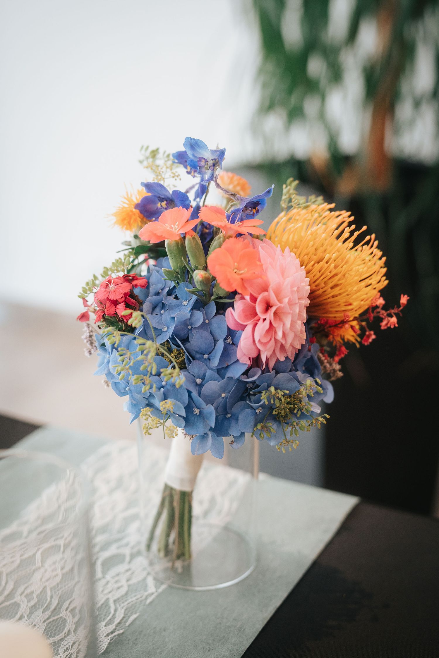 Colorful wedding bouquet of blue hydrangea, pink dahlia, orange blooms and yellow pincushion protea in a glass vase on a lace table runner.