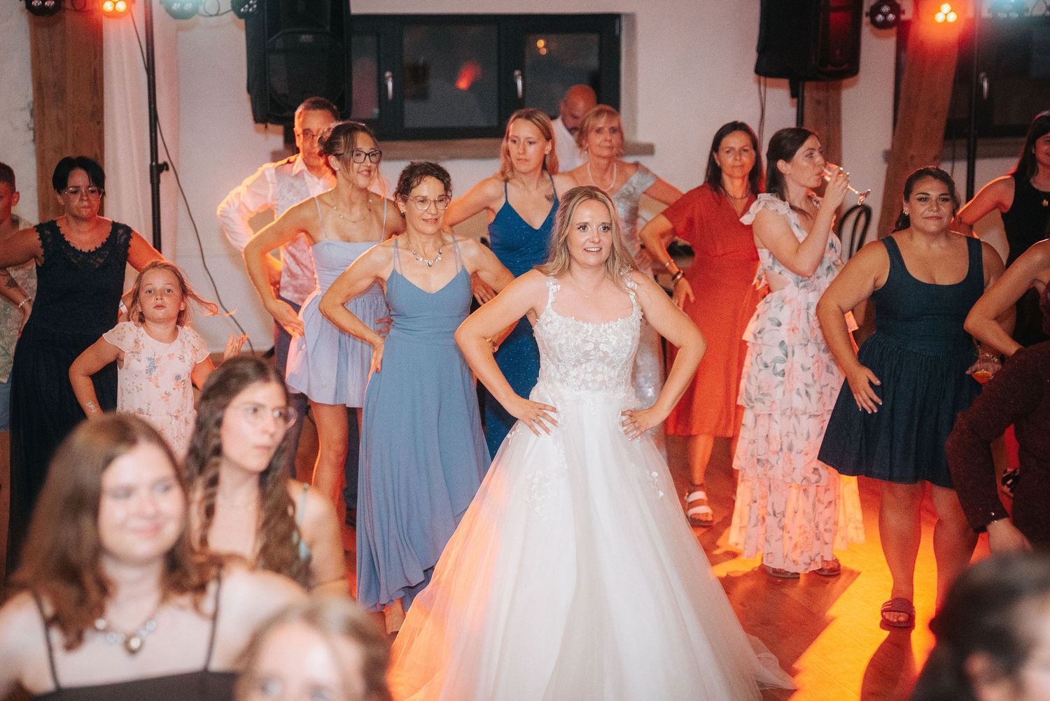 Bride in a white lace gown dances joyfully among smiling wedding guests in colorful dresses at an indoor reception.