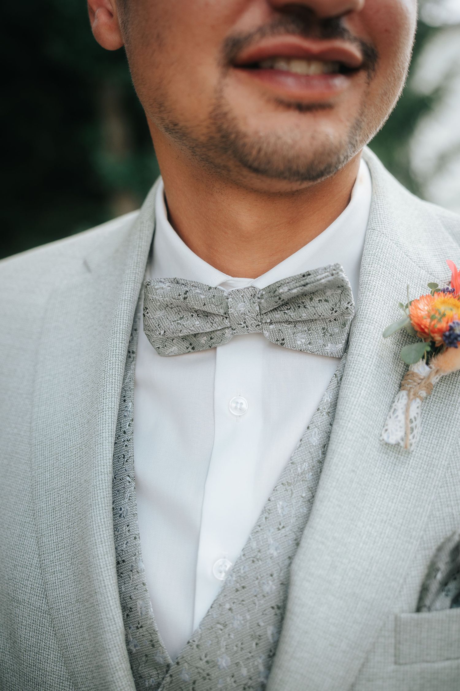 Smiling groom in a light gray suit, patterned gray bow tie and matching vest, with an orange floral boutonniere pinned to his lapel.