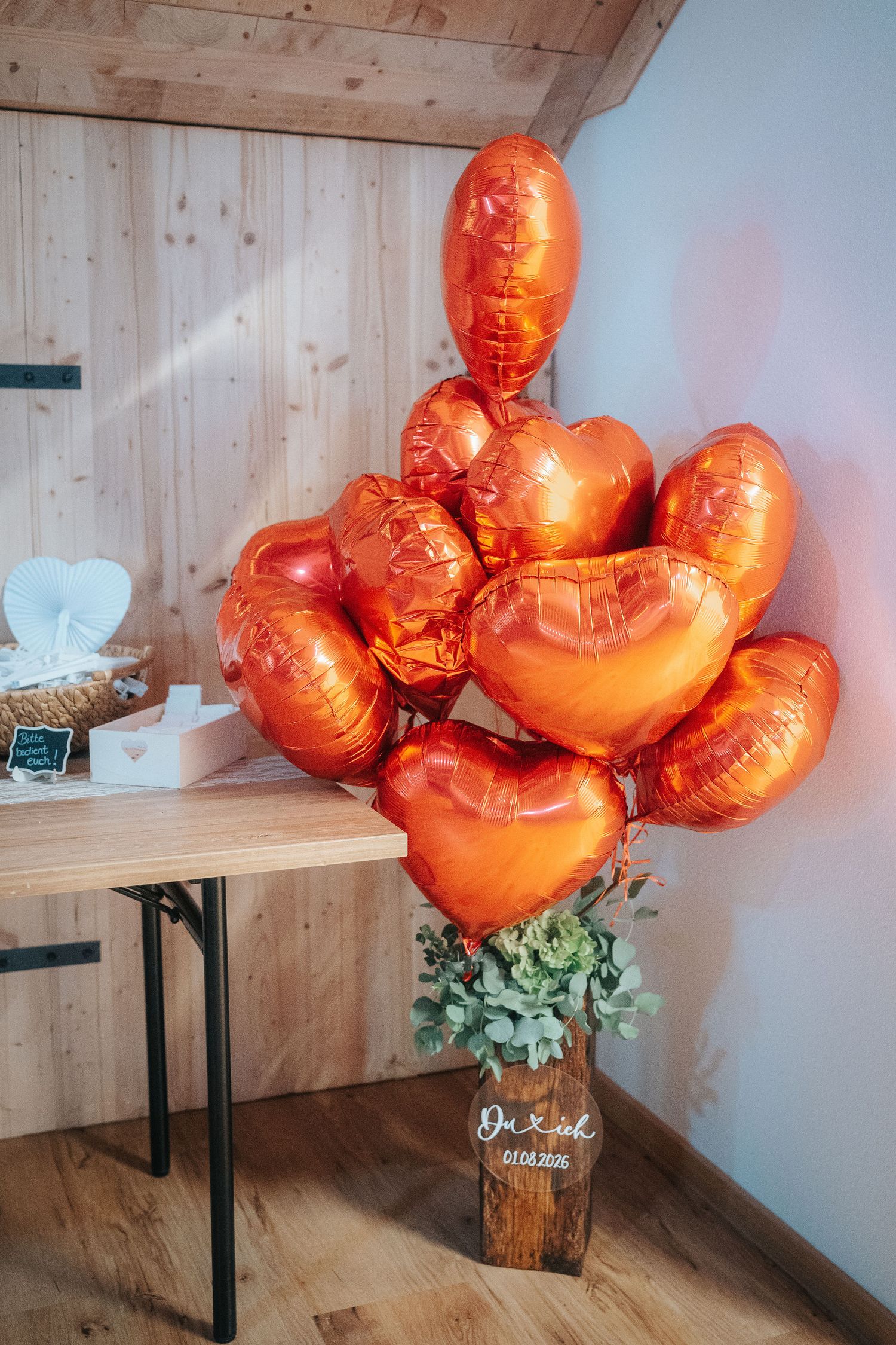 Orange heart-shaped foil balloons in a bouquet with eucalyptus greenery on a wooden stump beside a rustic table at a wedding venue.