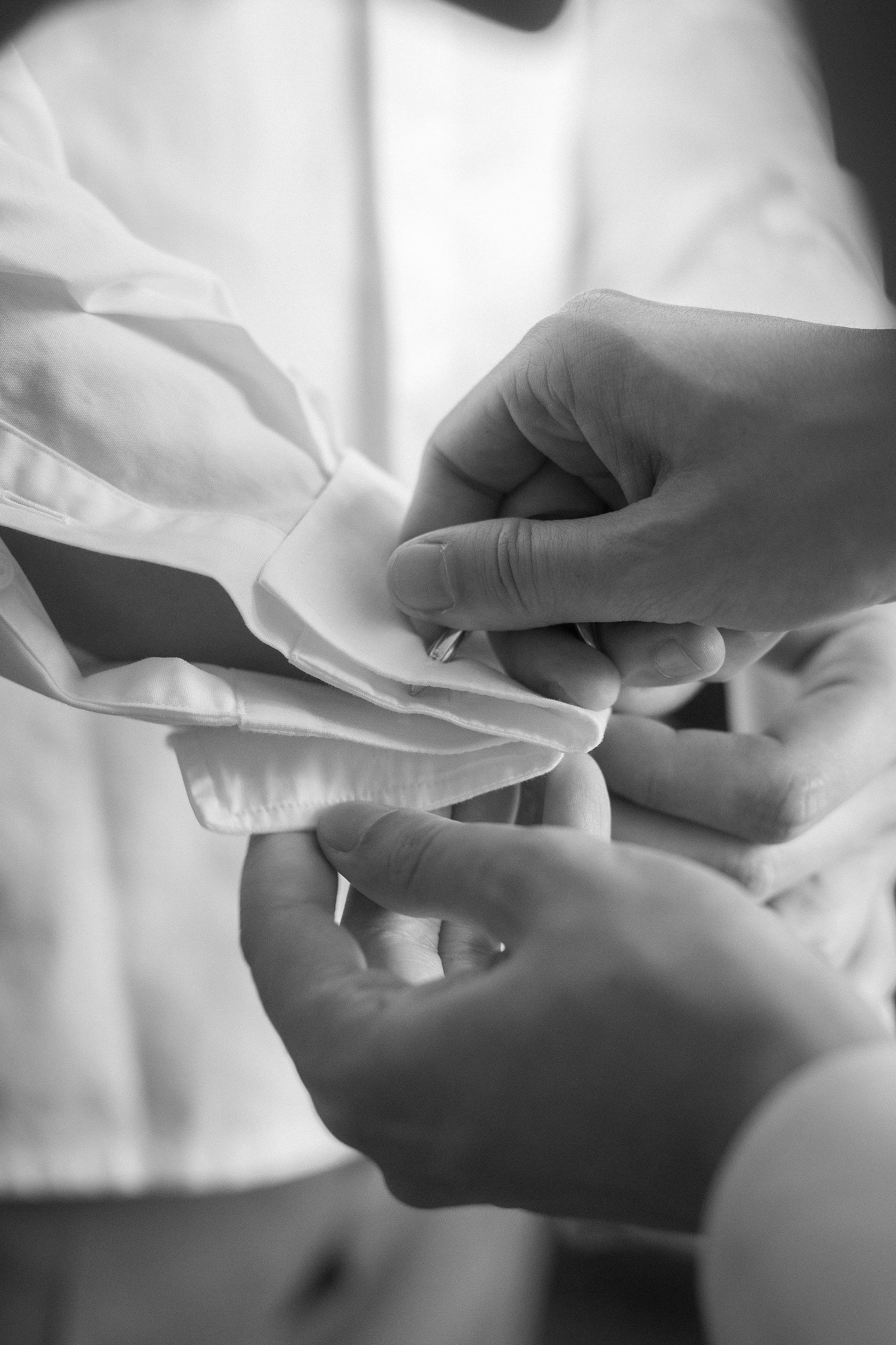 Black and white close-up of hands fastening a cufflink on a crisp white dress shirt cuff, a classic groom getting ready moment.