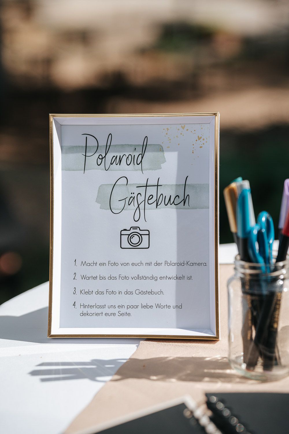 A framed sign with German text 'Polaroid Gastebuch' and instructions next to a glass jar with blue pens.