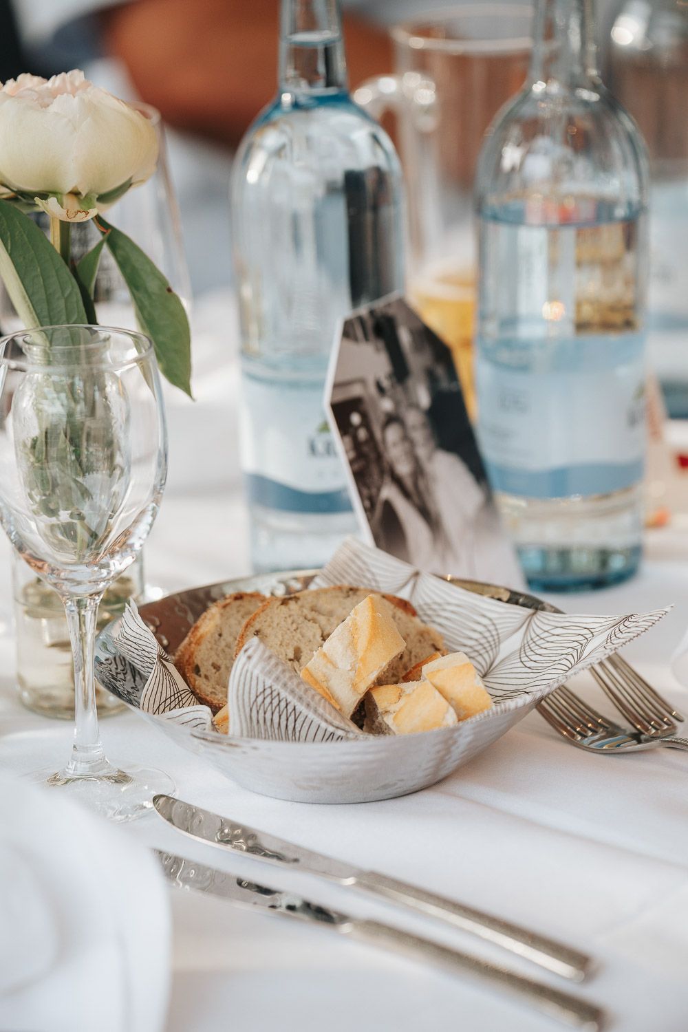 Elegant table setting with crusty bread basket, white peony in vase, water bottles and silverware on crisp white tablecloth.