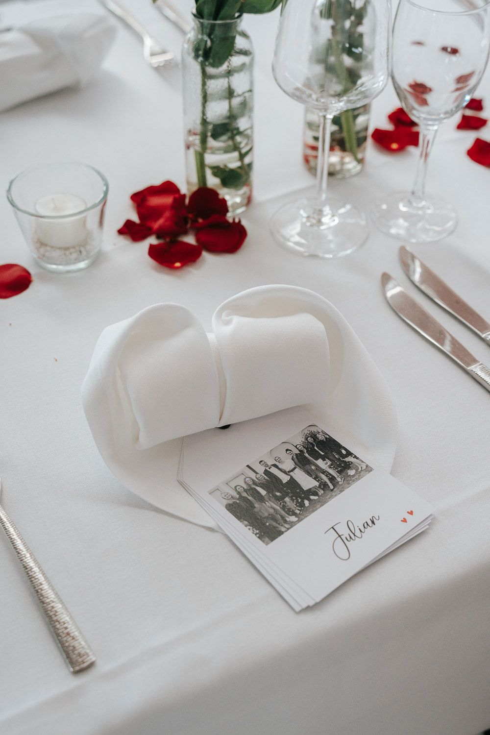 Elegant place card settings on a white tablecloth with red rose petals and glassware create a romantic wedding atmosphere.