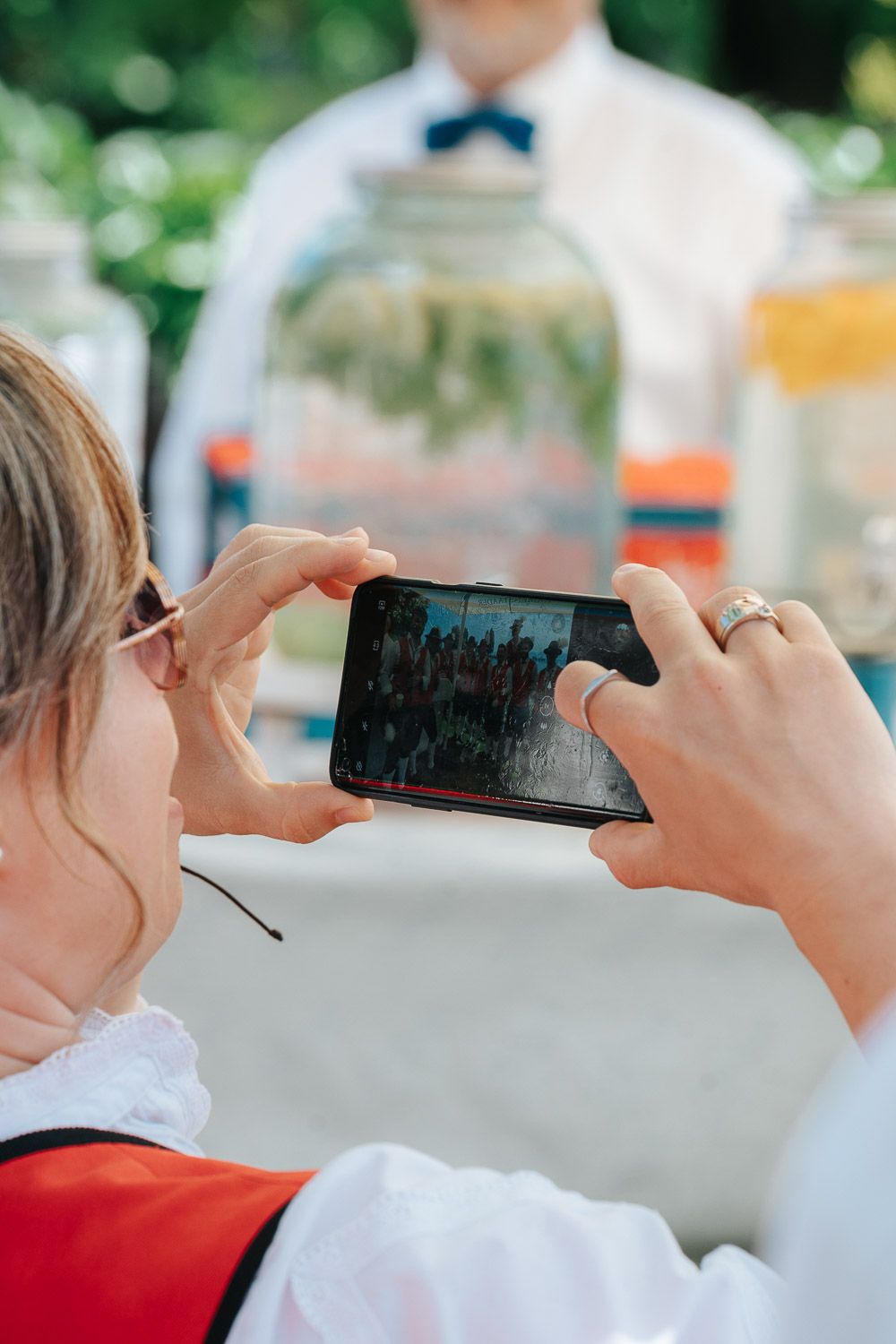 Person in red shirt taking a photo with a smartphone while sitting outdoors on a sunny day.