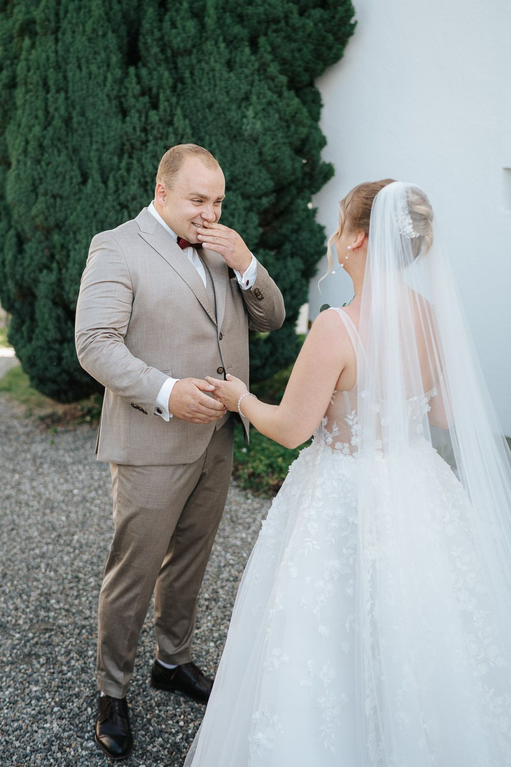 An emotional outdoor wedding moment captures a groom in beige suit and bride in white dress near evergreen trees.