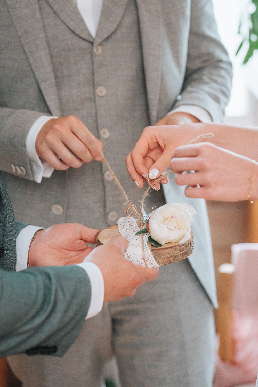 Wedding rings nestled in rustic burlap and lace holder being held between two people wearing gray suits.