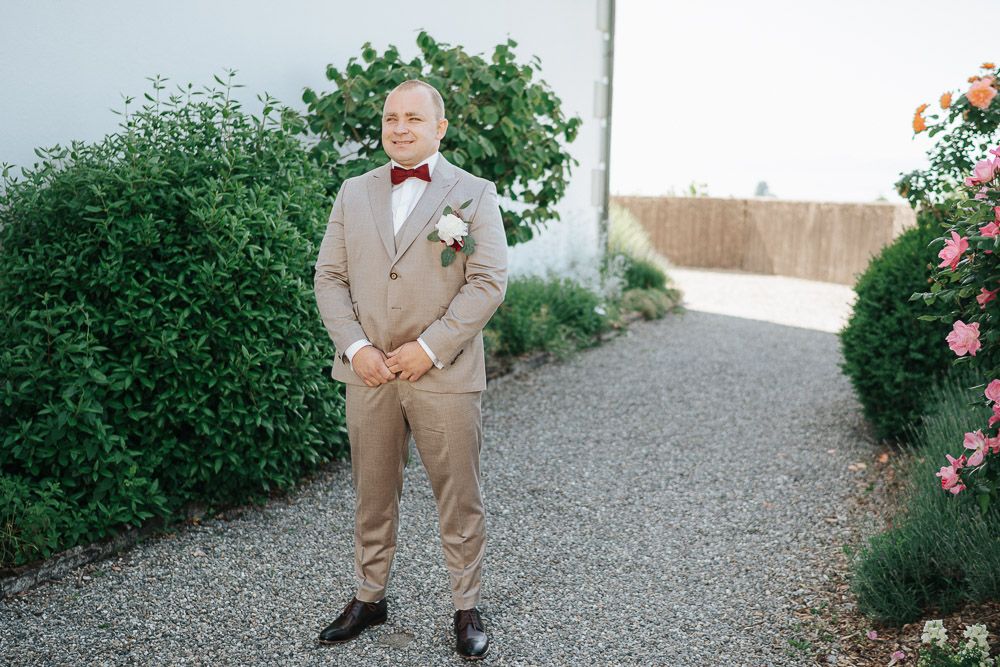 Groom in light tan suit and bow tie stands on a garden path surrounded by green shrubs and pink roses.