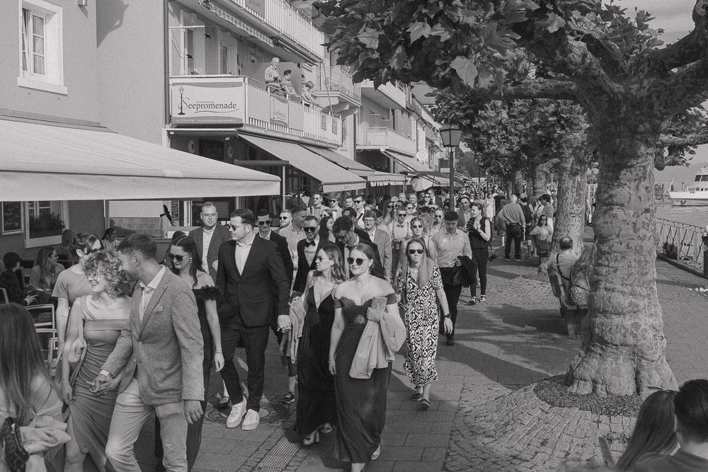 Vintage black and white photo of people walking down a busy sidewalk lined with shops and trees.