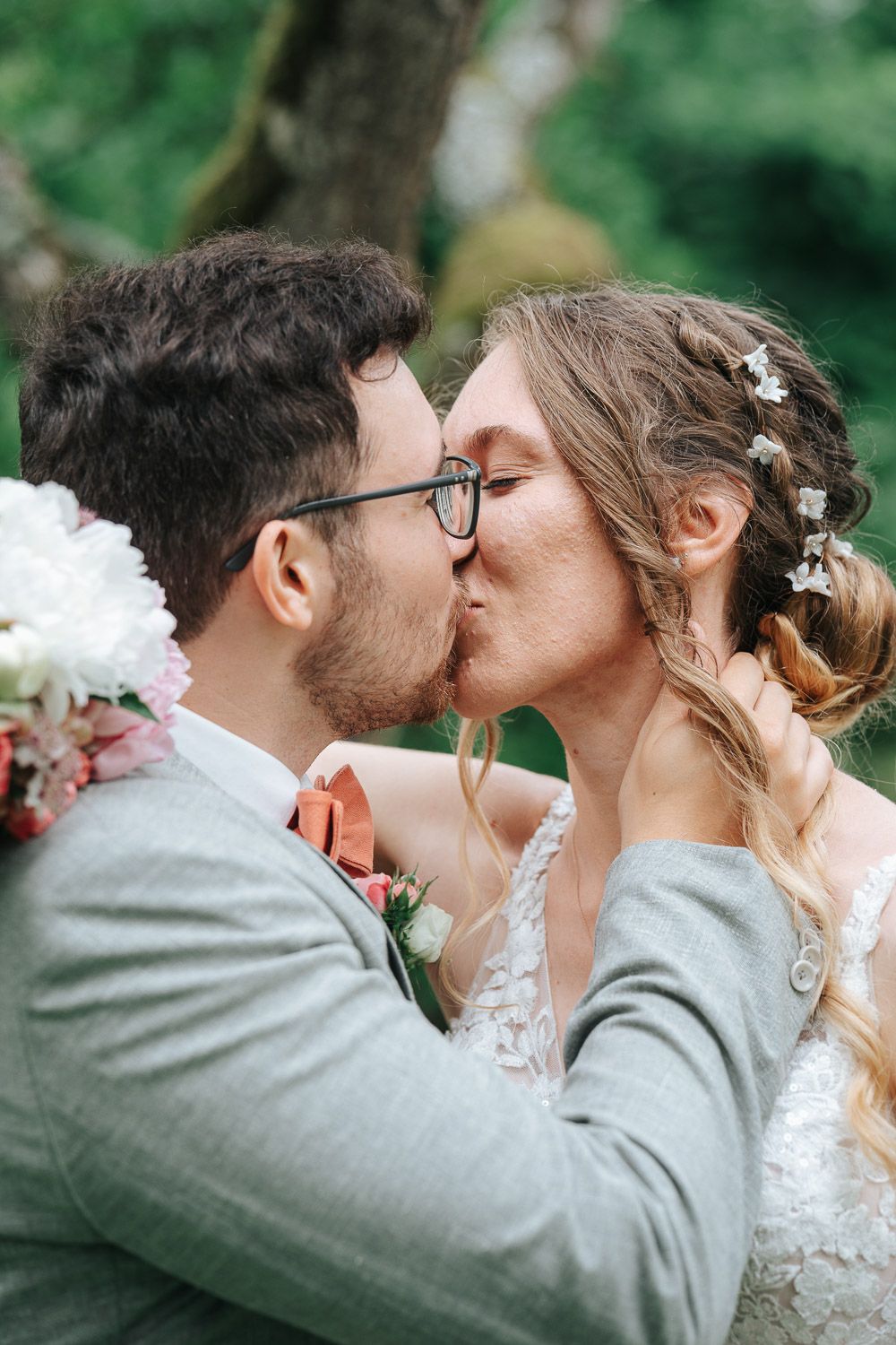A romantic wedding kiss captured outdoors with white flowers and delicate hair accessories against a natural green backdrop.