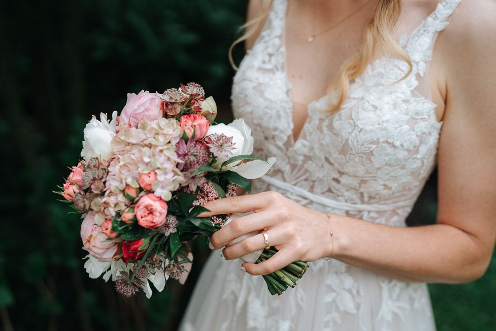 A delicate pink and white bridal bouquet held against a lace wedding dress with detailed floral embellishments.