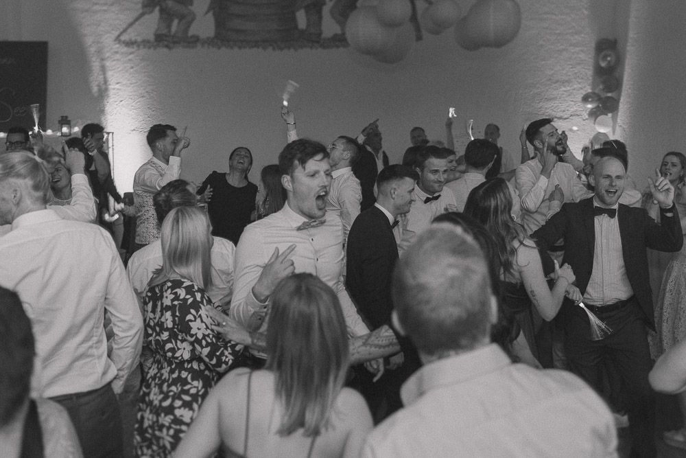 Black and white photo of a crowded party or wedding reception with people dancing and mingling at an indoor venue.