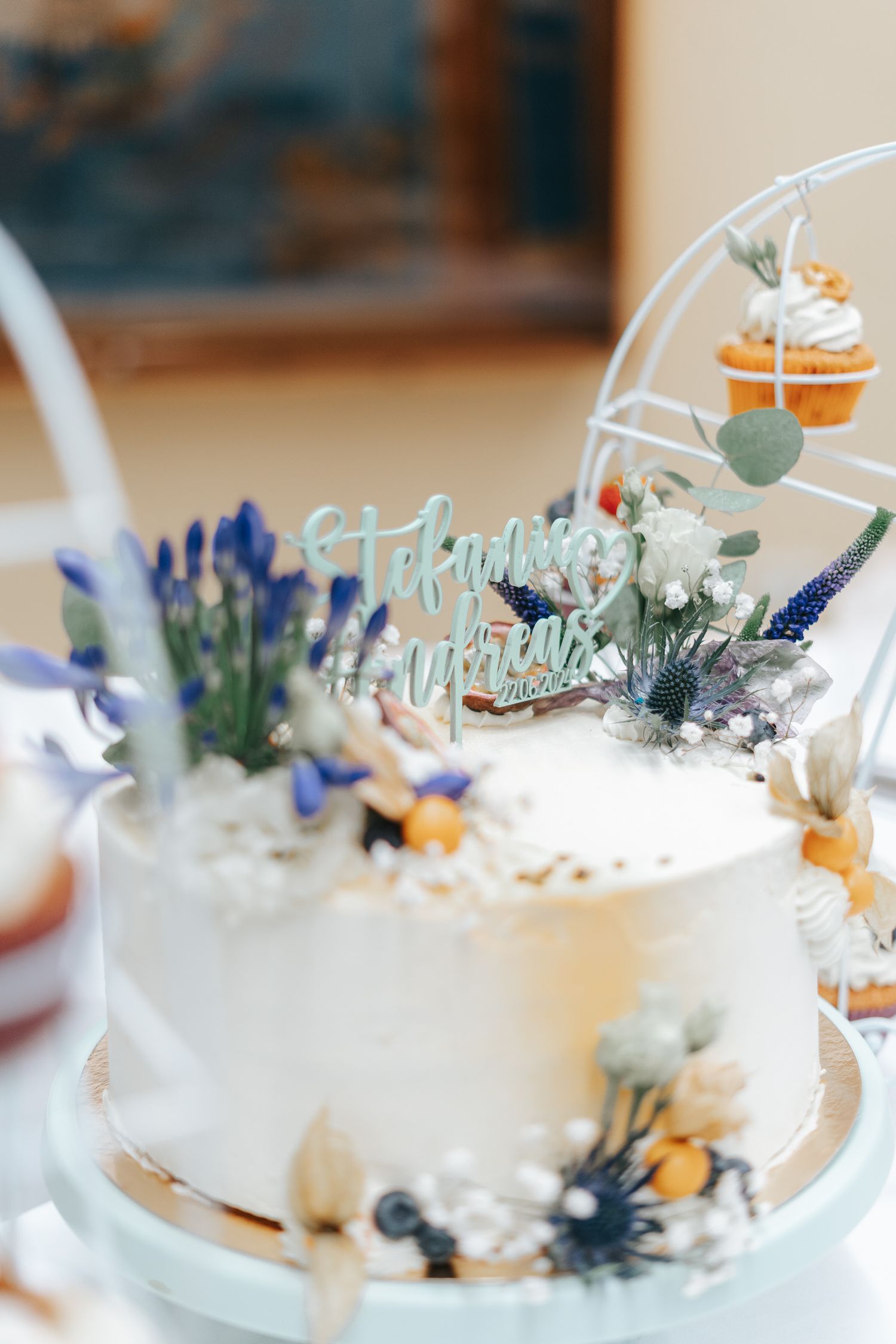White wedding cake decorated with blue lavender flowers, eucalyptus leaves and a gold 'Happy Anniversary' cake topper.