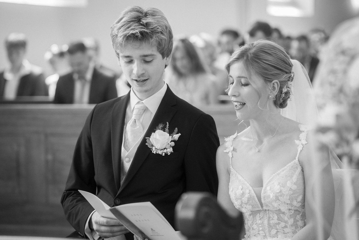 Black and white photo of a bride and groom reading from a booklet during a wedding ceremony, with guests seated in pews behind them.