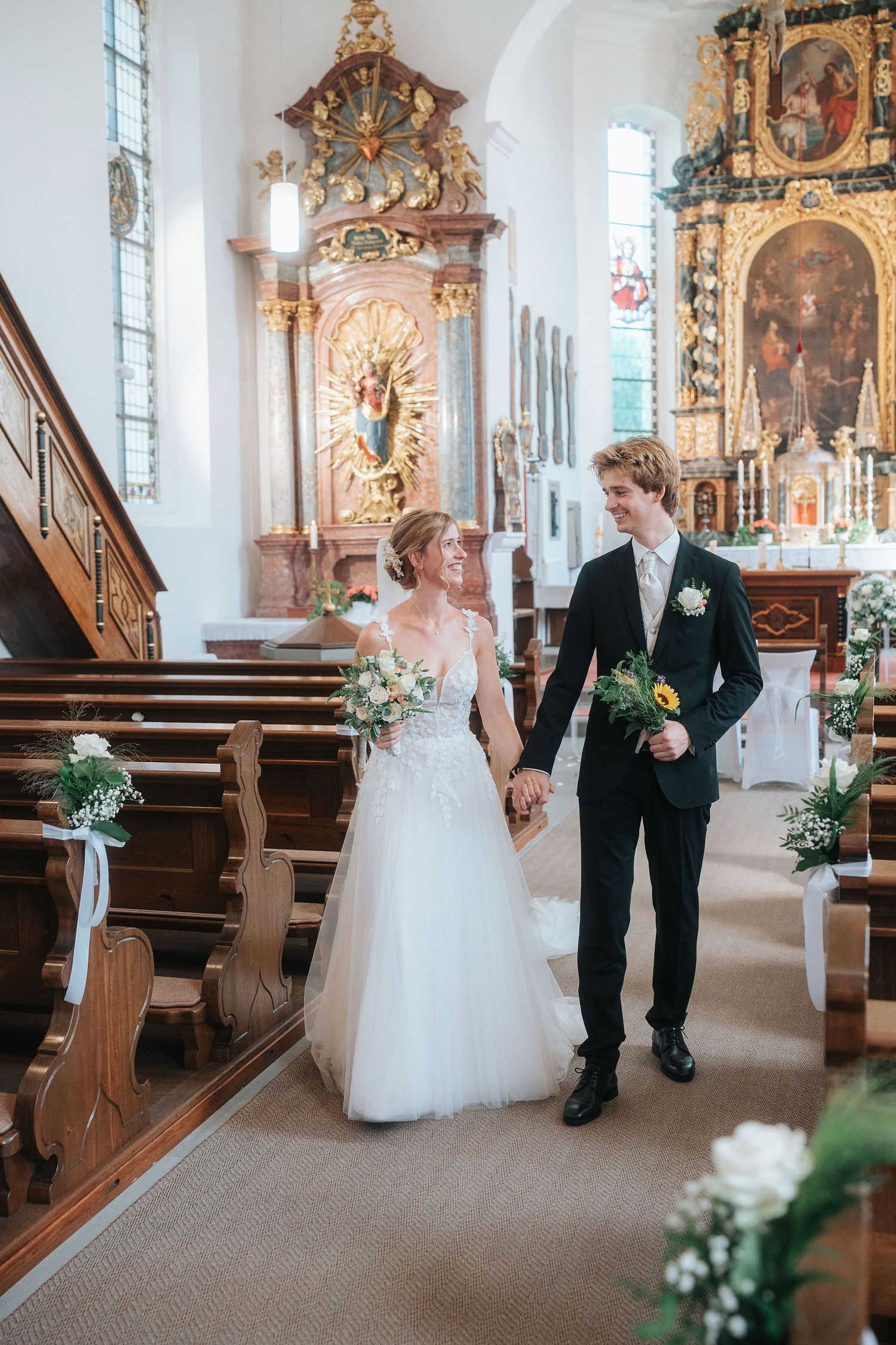 Bride and groom walking hand in hand down the aisle of an ornate church after their wedding ceremony.
