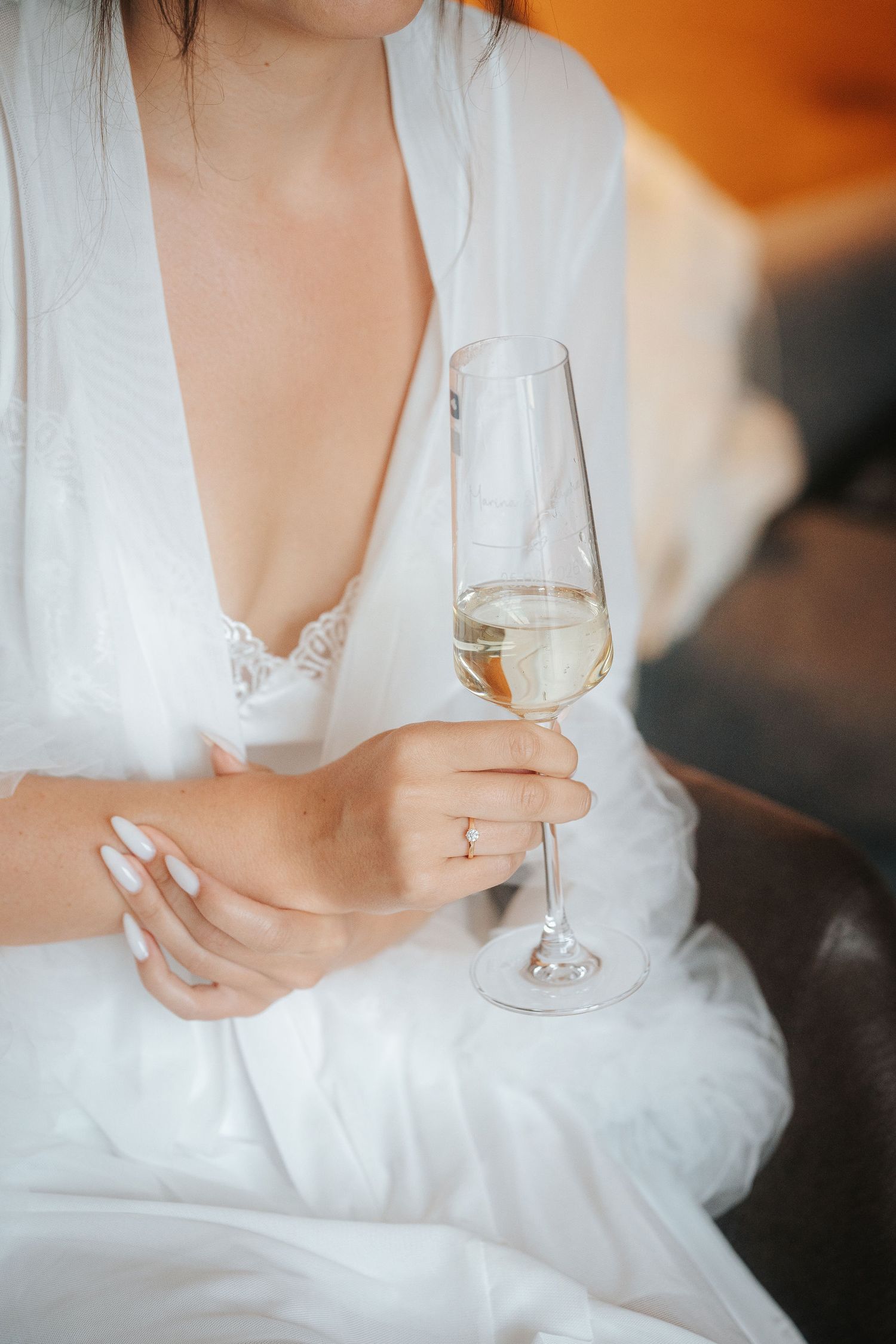 Bride in a white satin robe with lace trim holds a glass of champagne while sitting in an armchair, wearing a ring and white manicure.