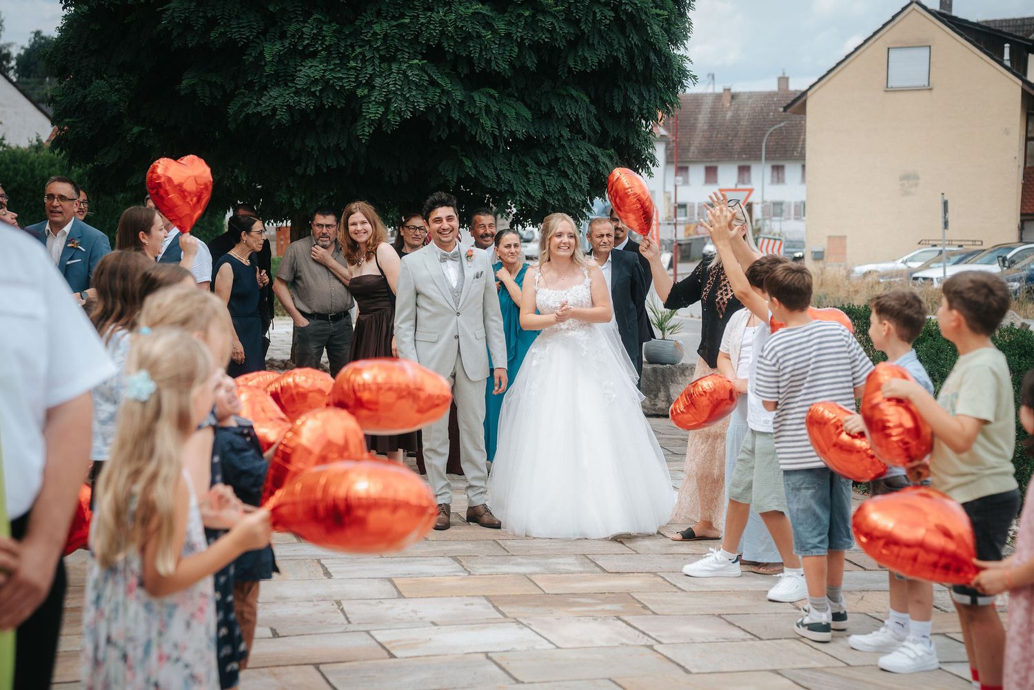 Bride and groom walk past cheering wedding guests holding red heart-shaped balloons on a sunny outdoor cobblestone square.