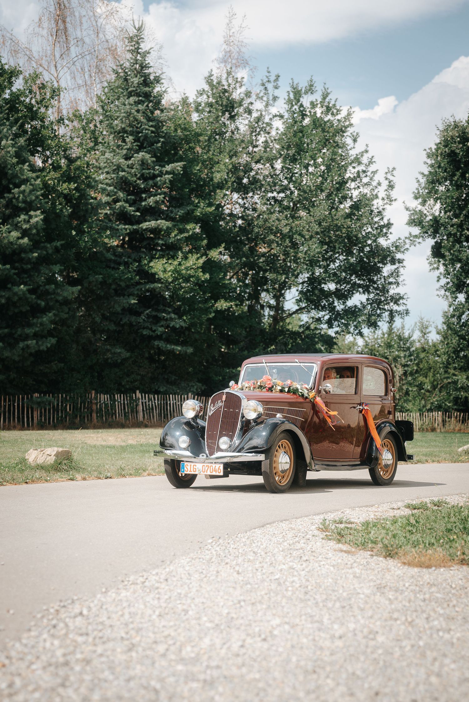 Vintage brown wedding car decorated with a floral arrangement parked on a paved country road beside green evergreen trees.