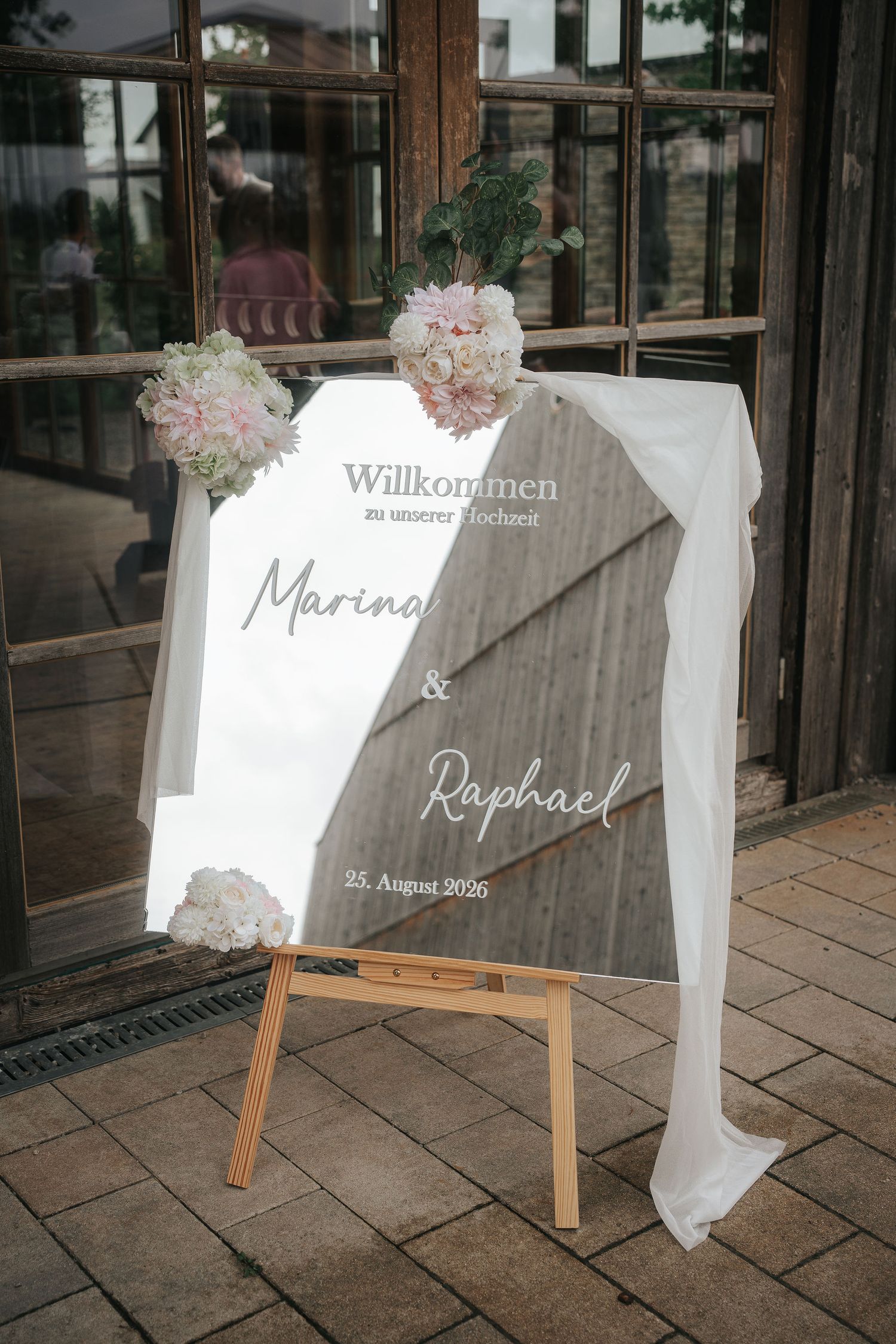 Mirrored wedding welcome sign for Marina and Raphael dated 25 August 2026, on a wooden easel with white fabric and pink floral arrangements.