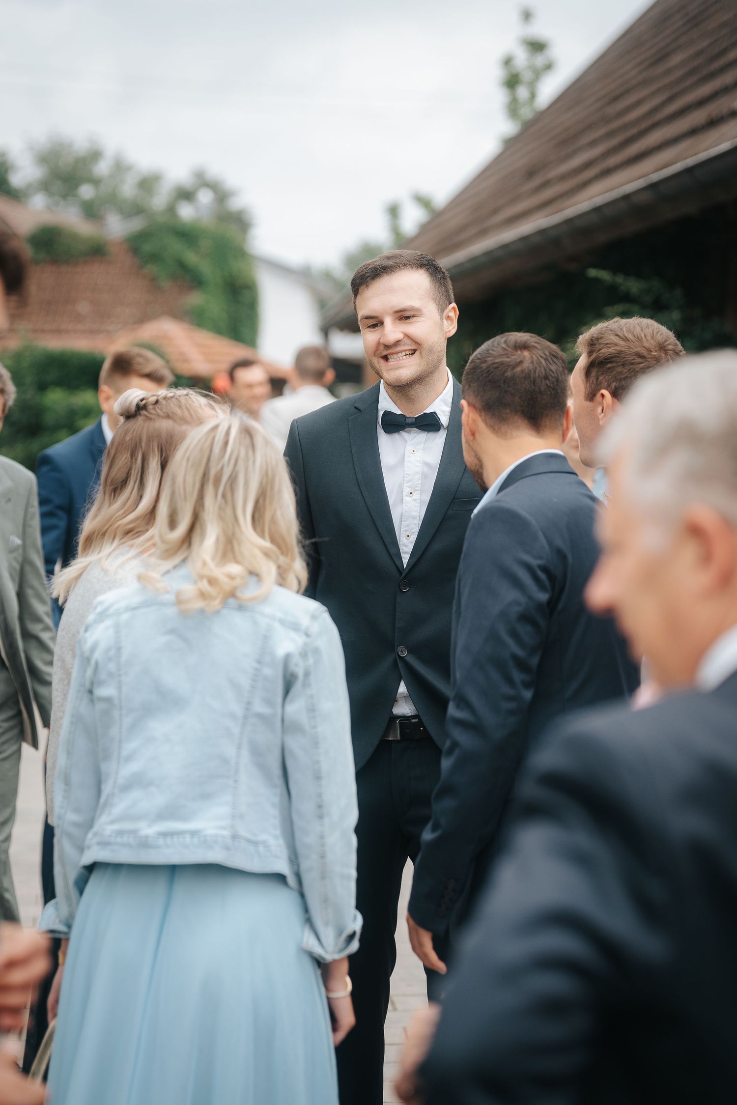 Smiling groom in a navy suit and bow tie chats with wedding guests outdoors, including a woman in a denim jacket and light blue skirt.