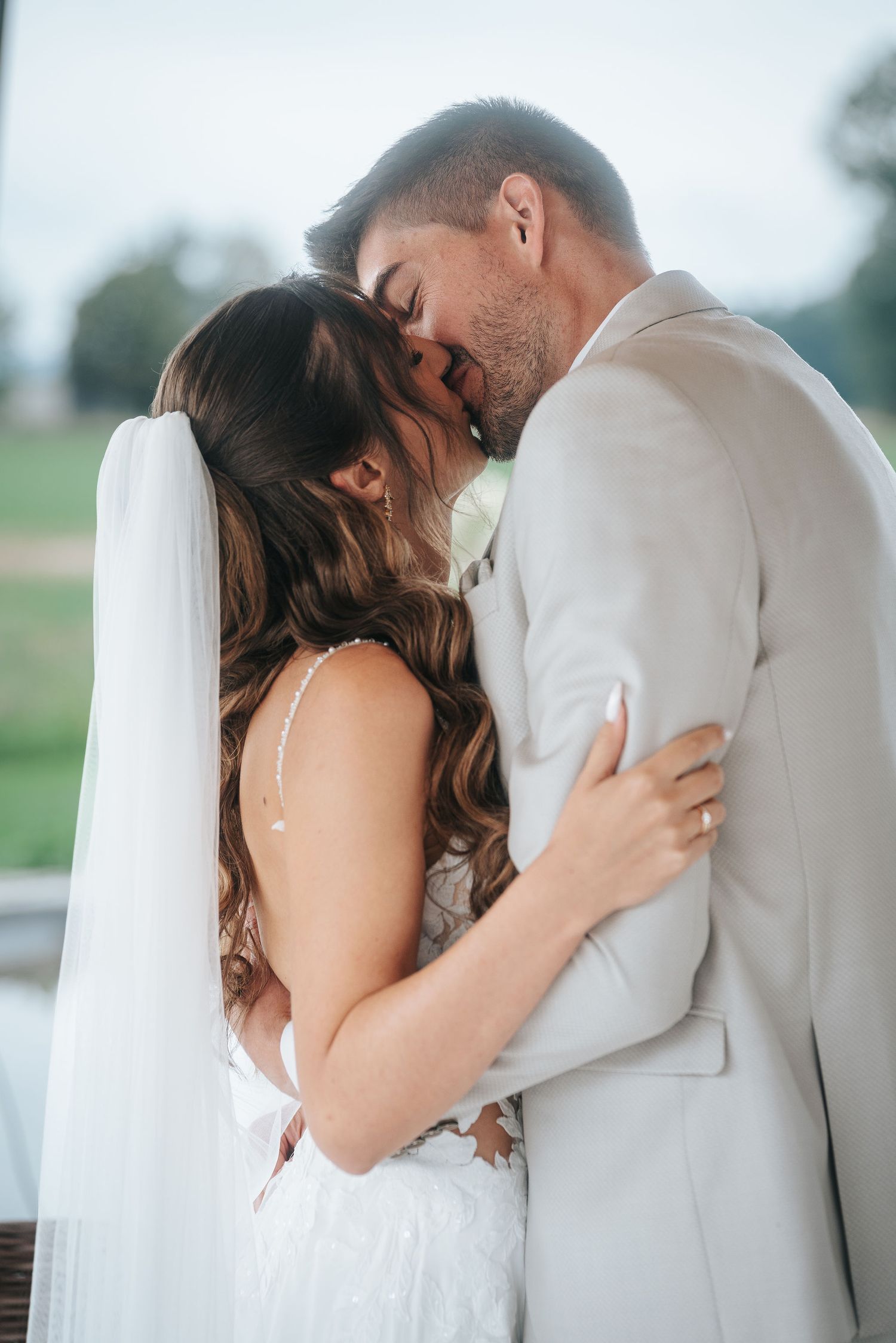 Bride in a white lace gown and long veil kisses her groom in a light gray suit during a romantic outdoor wedding moment.