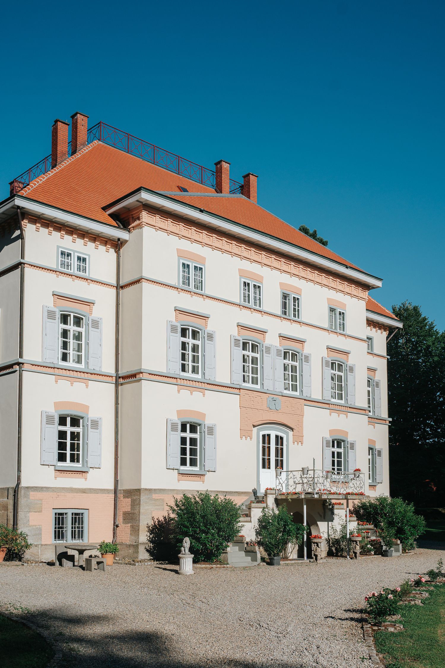 Elegant cream-colored manor house with orange tile roof, gray shutters, and a gravel courtyard with lush shrubs under a clear blue sky.