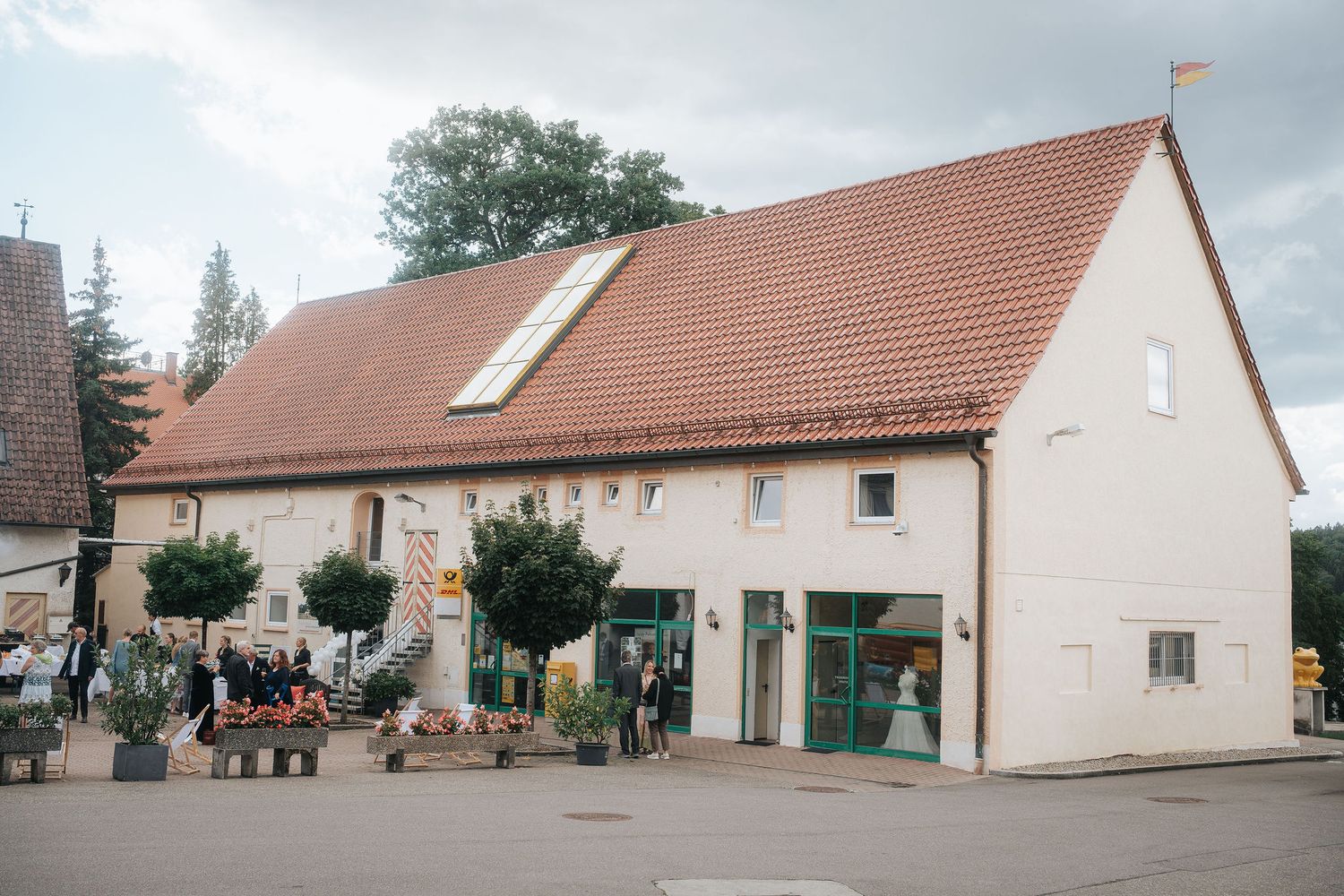 Rustic two-story building with a red tile roof, skylight and large glass doors, with guests gathered outside on a cobblestone square.
