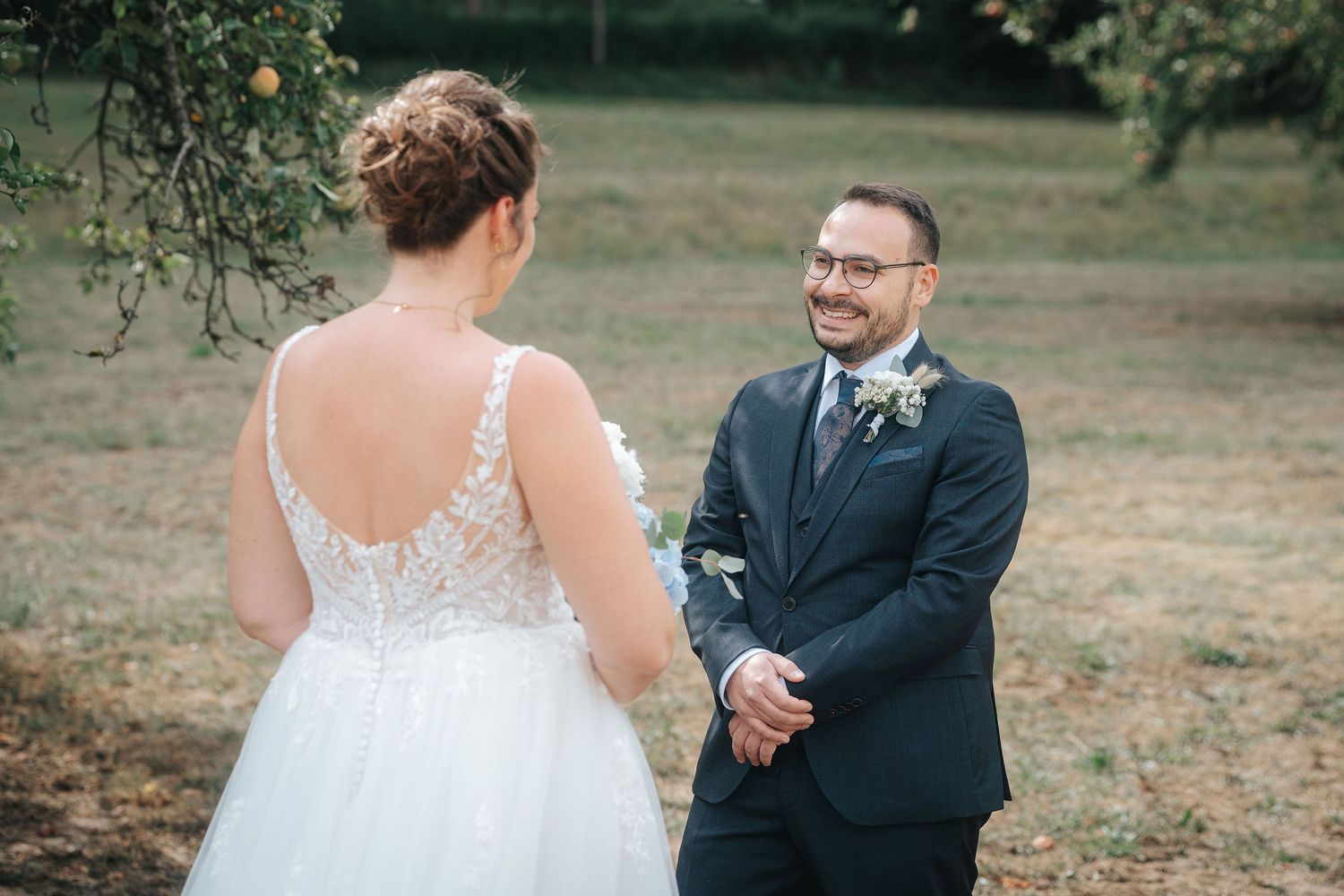 Smiling groom in a navy suit with a boutonniere looks at his bride in a lace-back wedding dress during an outdoor first look.