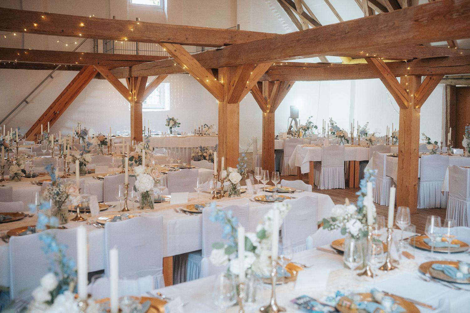 Rustic barn wedding reception hall with exposed timber beams, long tables with white linens, candles, greenery and floral centerpieces.