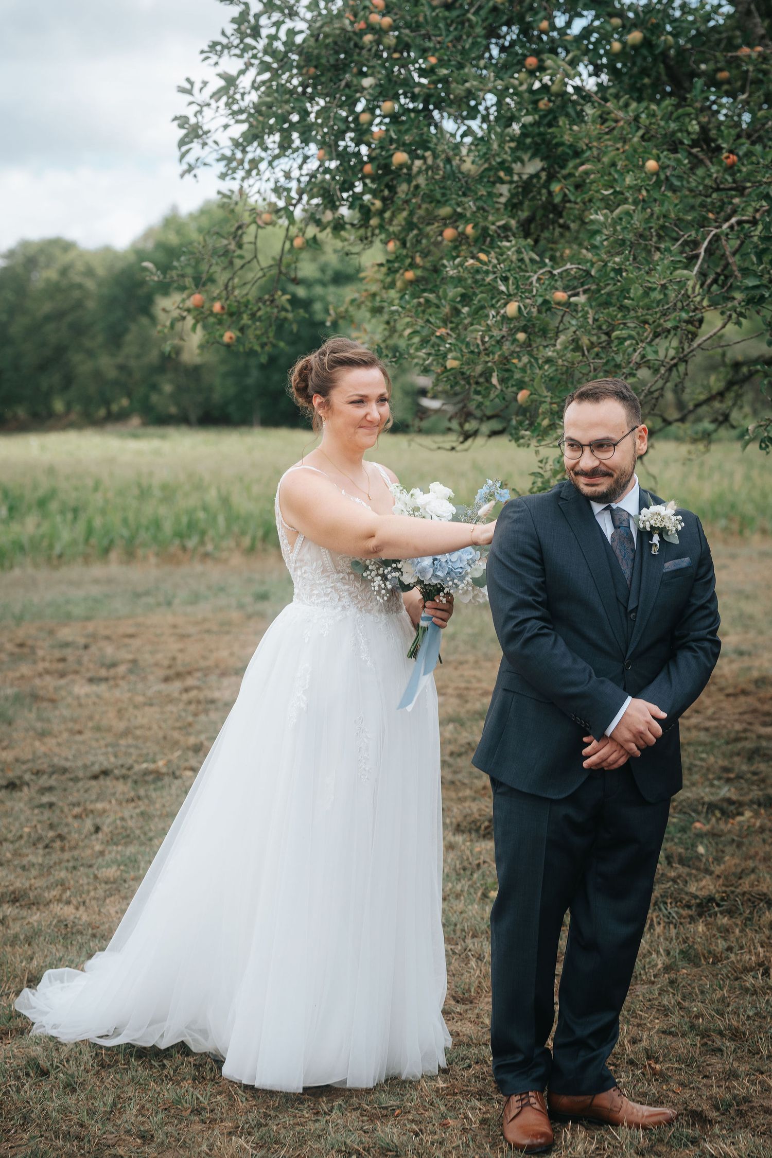 Bride in a white tulle gown holding a blue bouquet reaches toward a smiling groom in a navy suit during an outdoor orchard wedding.
