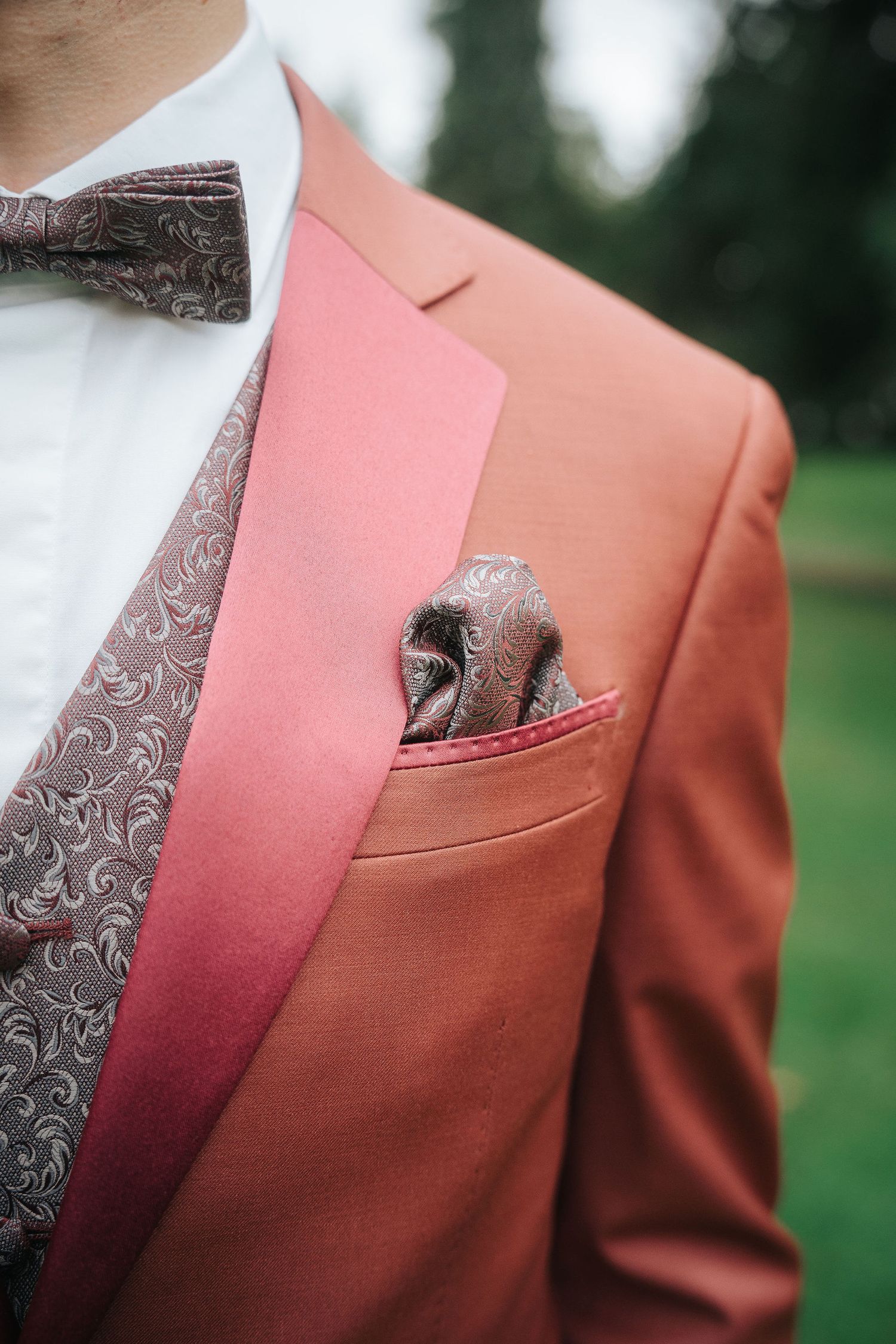 Groom in a terracotta suit jacket with satin pink lapel, paisley bow tie, matching pocket square and patterned vest outdoors.