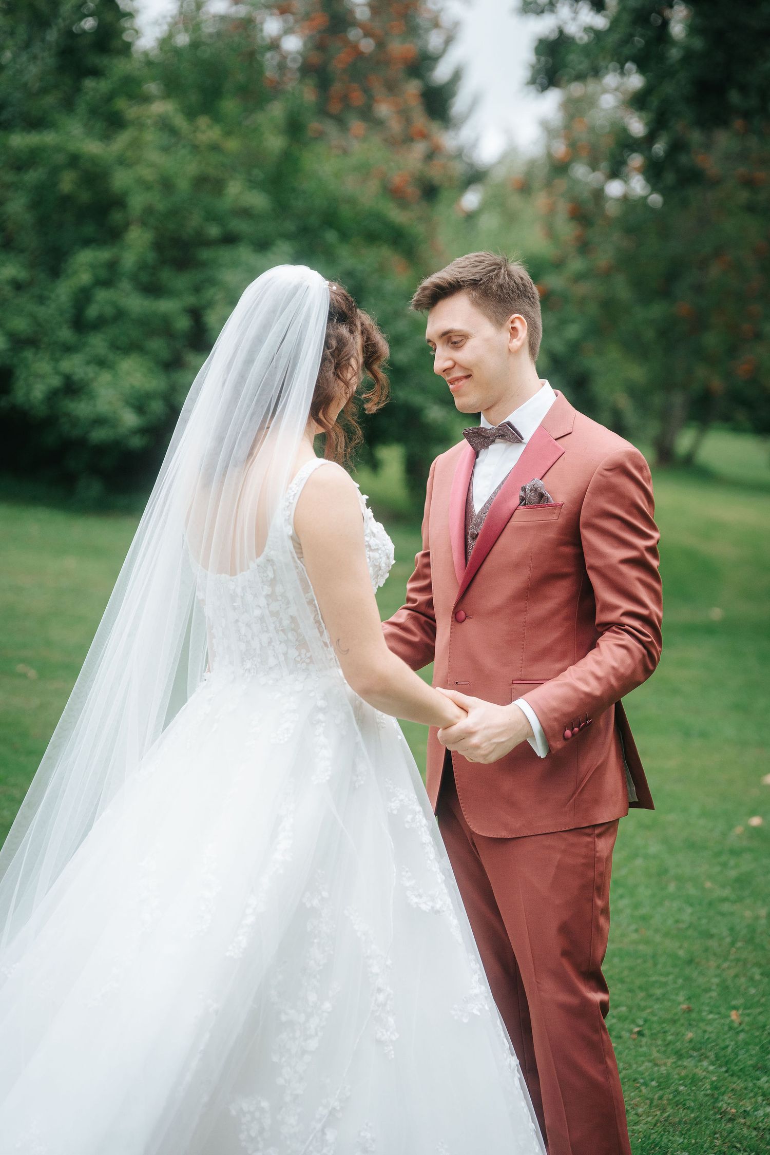 Bride in a white lace gown and long veil holds hands with a smiling groom in a rust-colored suit and bow tie on a green lawn.