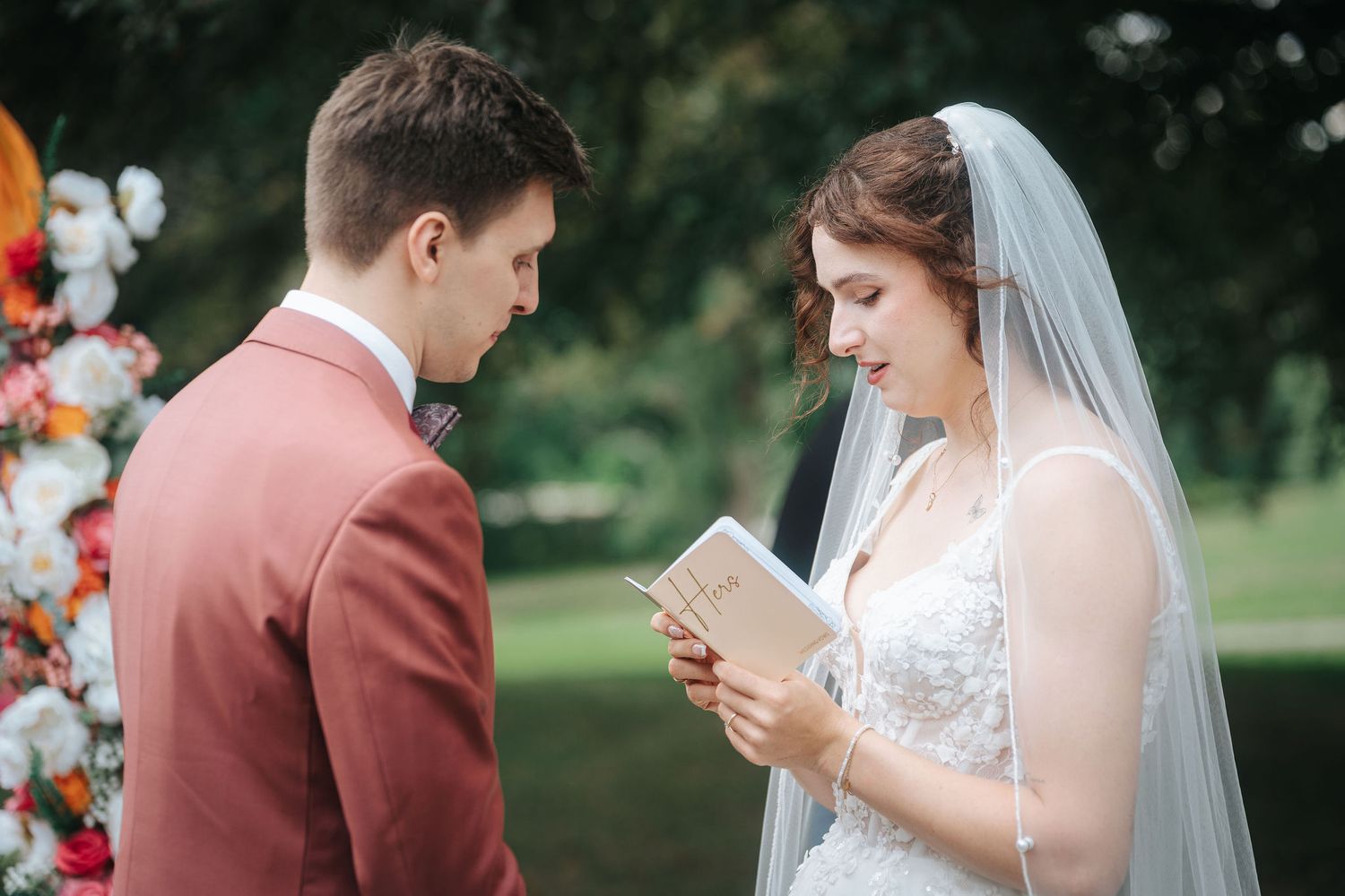 Bride in a lace gown and veil reads her handwritten vows from a small book to a groom in a rust suit during an outdoor wedding ceremony.