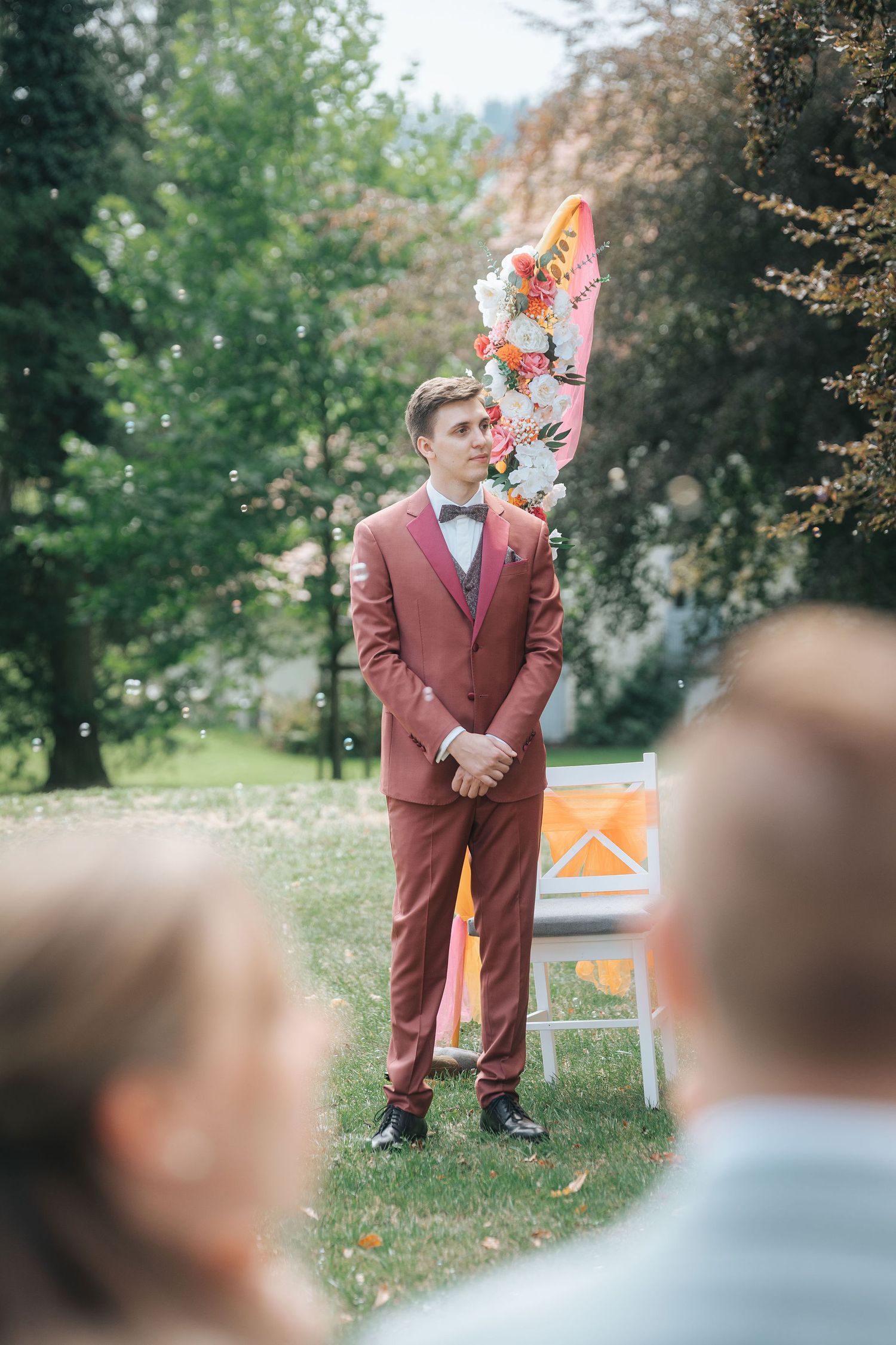 Groom in a rust-colored suit and bow tie stands in an outdoor garden ceremony beside a floral arch as guests look on.