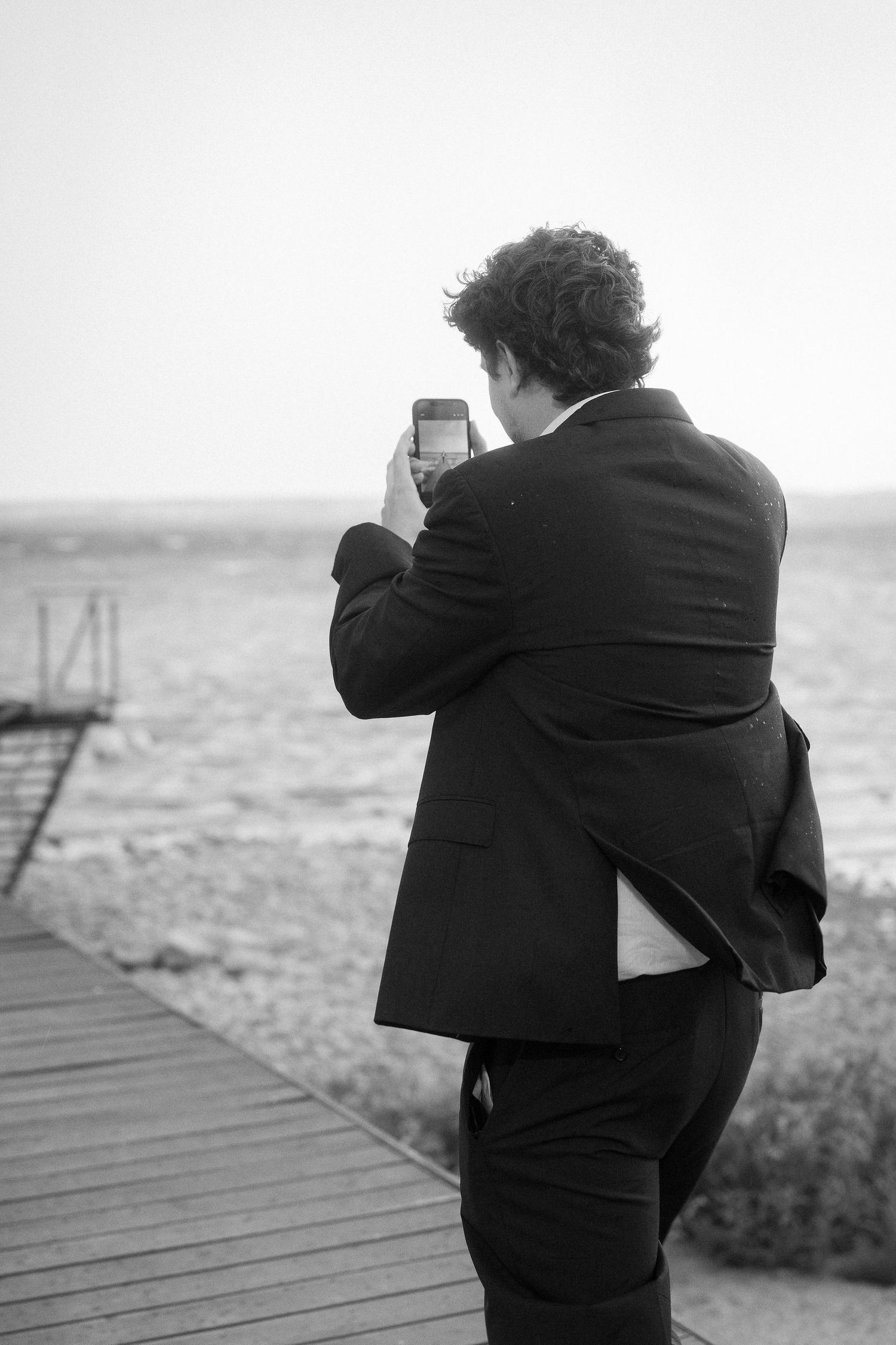 Black and white photo of a curly-haired man in a dark suit taking a picture with his smartphone on a wooden dock overlooking the water.