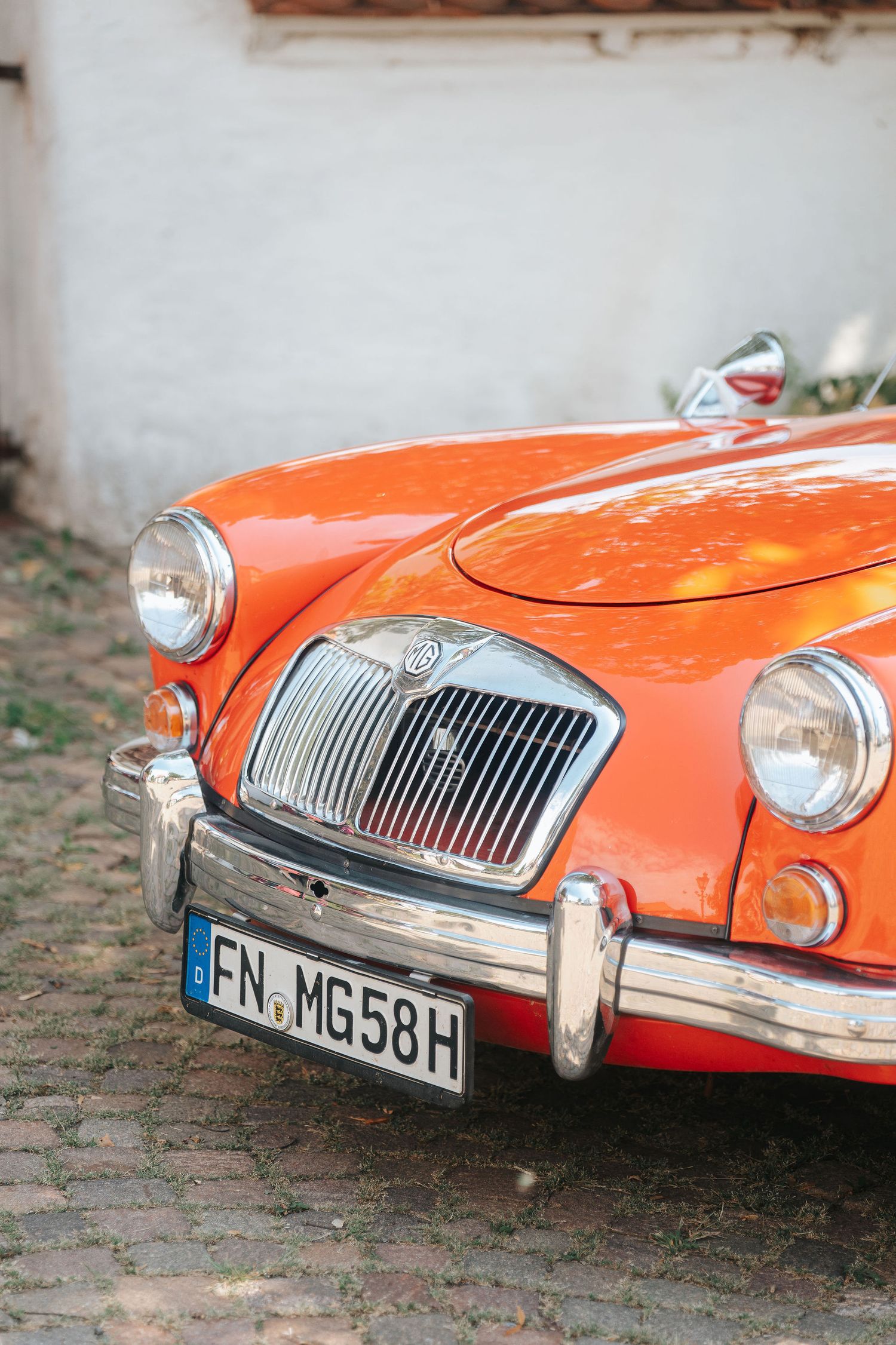 Front view of a vintage orange MG A roadster with chrome grille and bumper, parked on a cobblestone street beside a white wall.