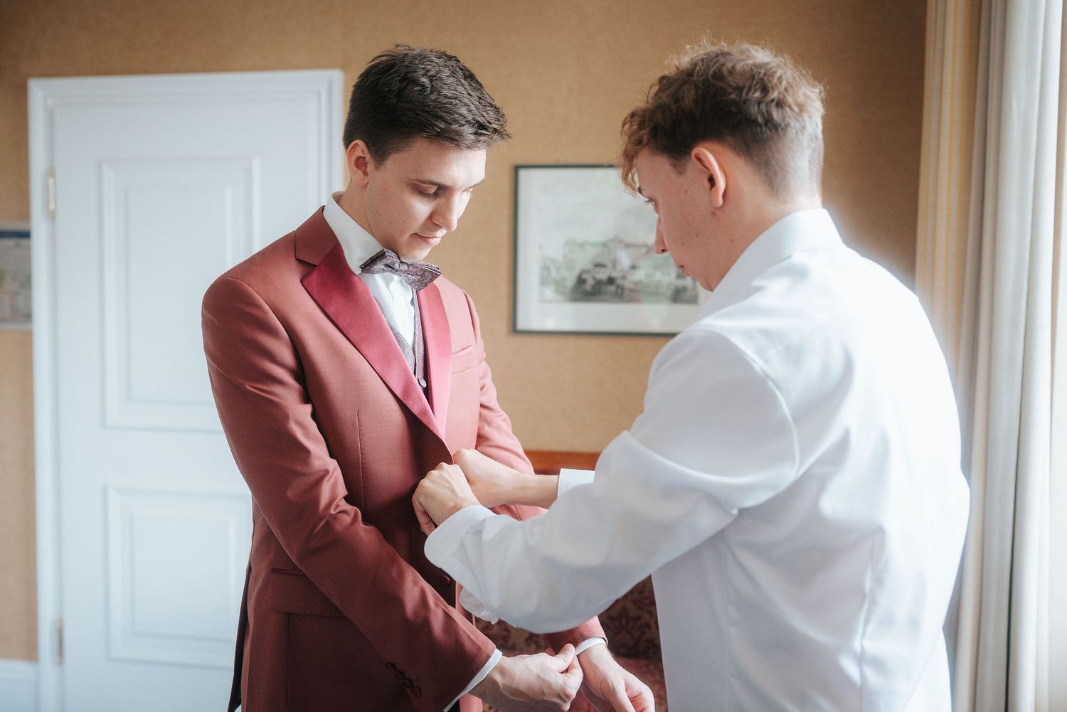 Groom in a burgundy suit and bow tie gets help adjusting his cuff from a man in a white shirt while getting ready for the wedding.