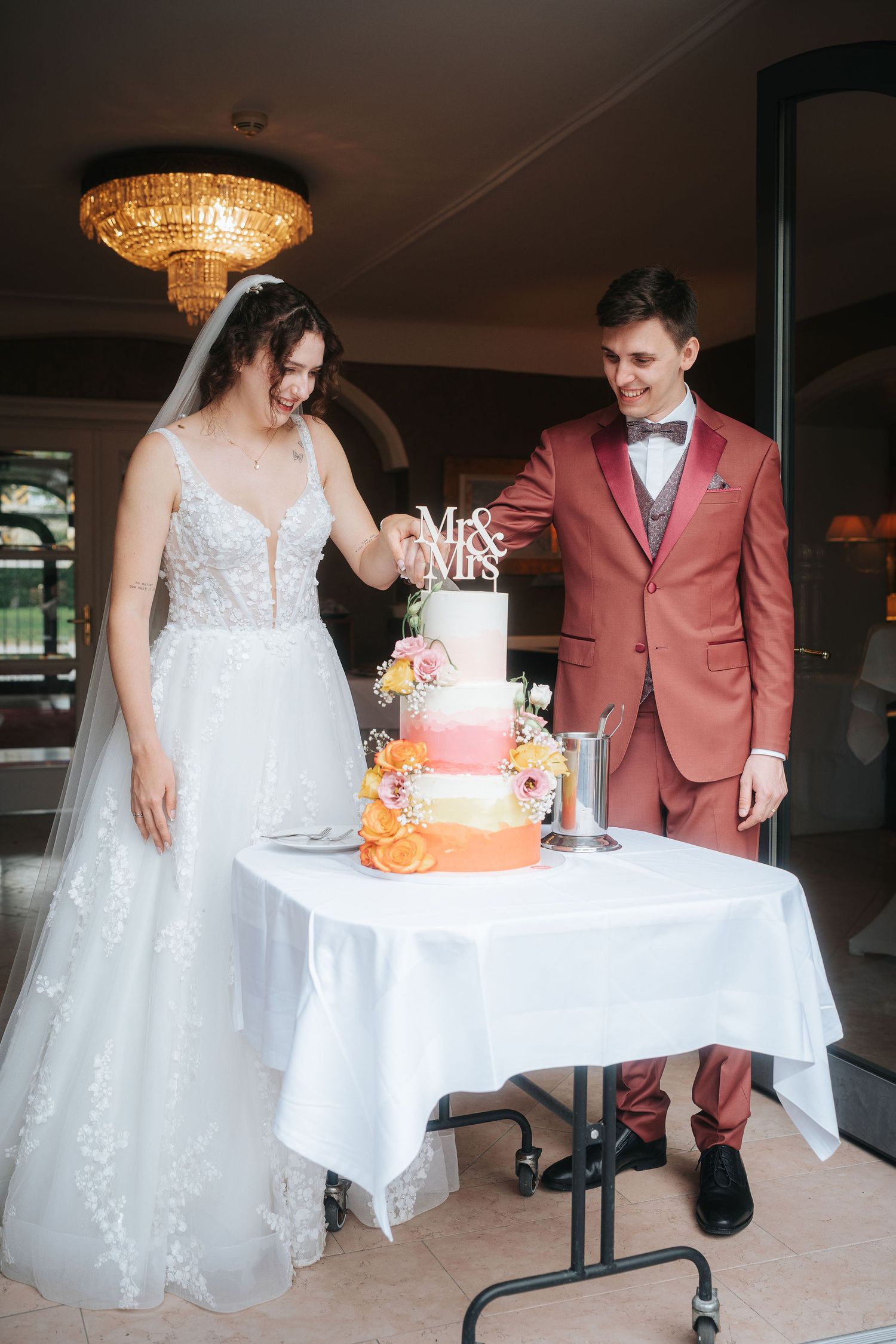 Smiling bride in a lace wedding gown and groom in a coral suit cut a colorful floral tiered Mr & Mrs wedding cake together.