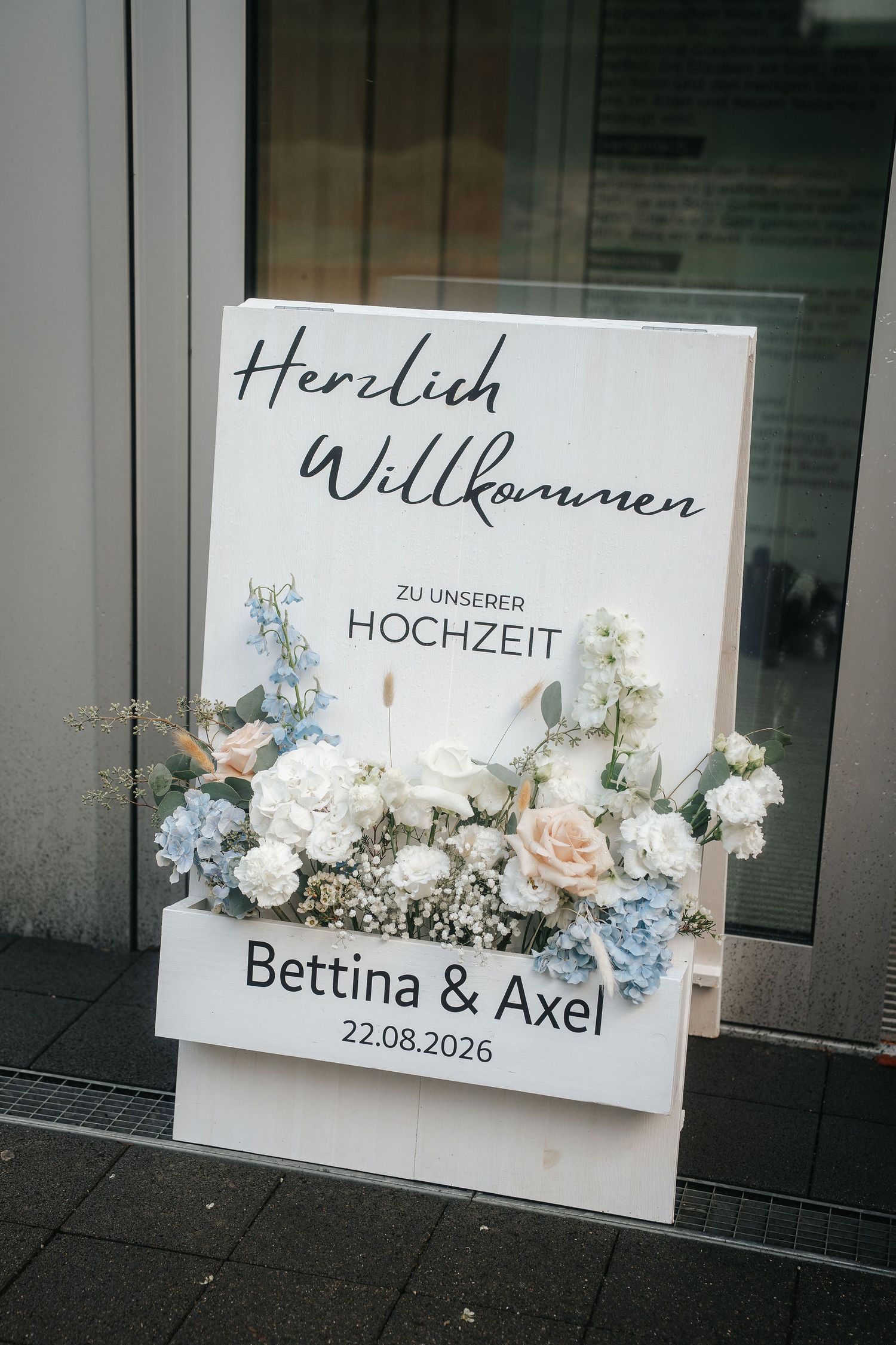 Wooden German wedding welcome sign for Bettina & Axel dated 22.08.2026, decorated with blue, white and peach flowers outside a glass door.