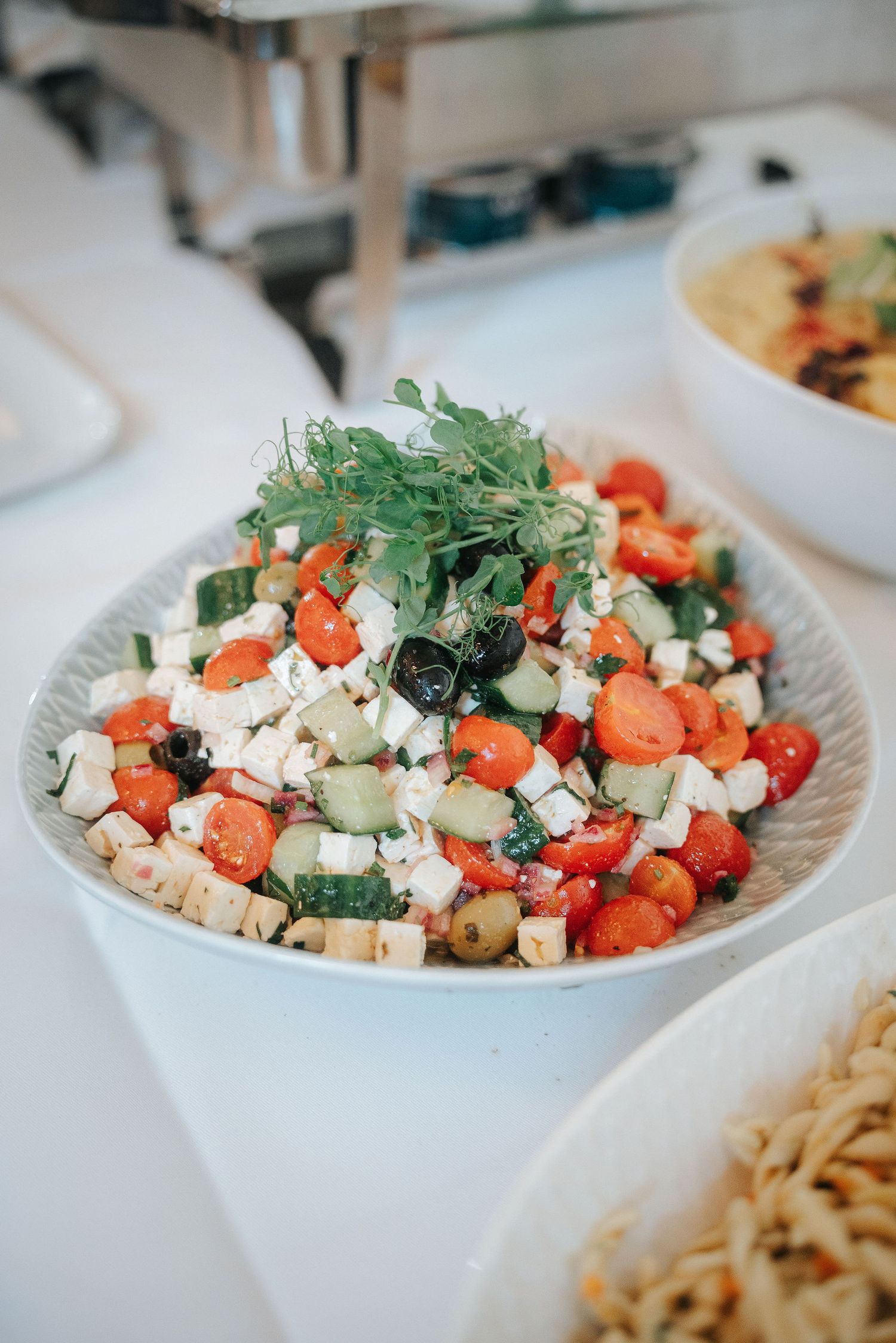 Fresh Greek salad with cherry tomatoes, cucumber, feta cheese, olives and herbs in a white bowl on a buffet table beside pasta salad.