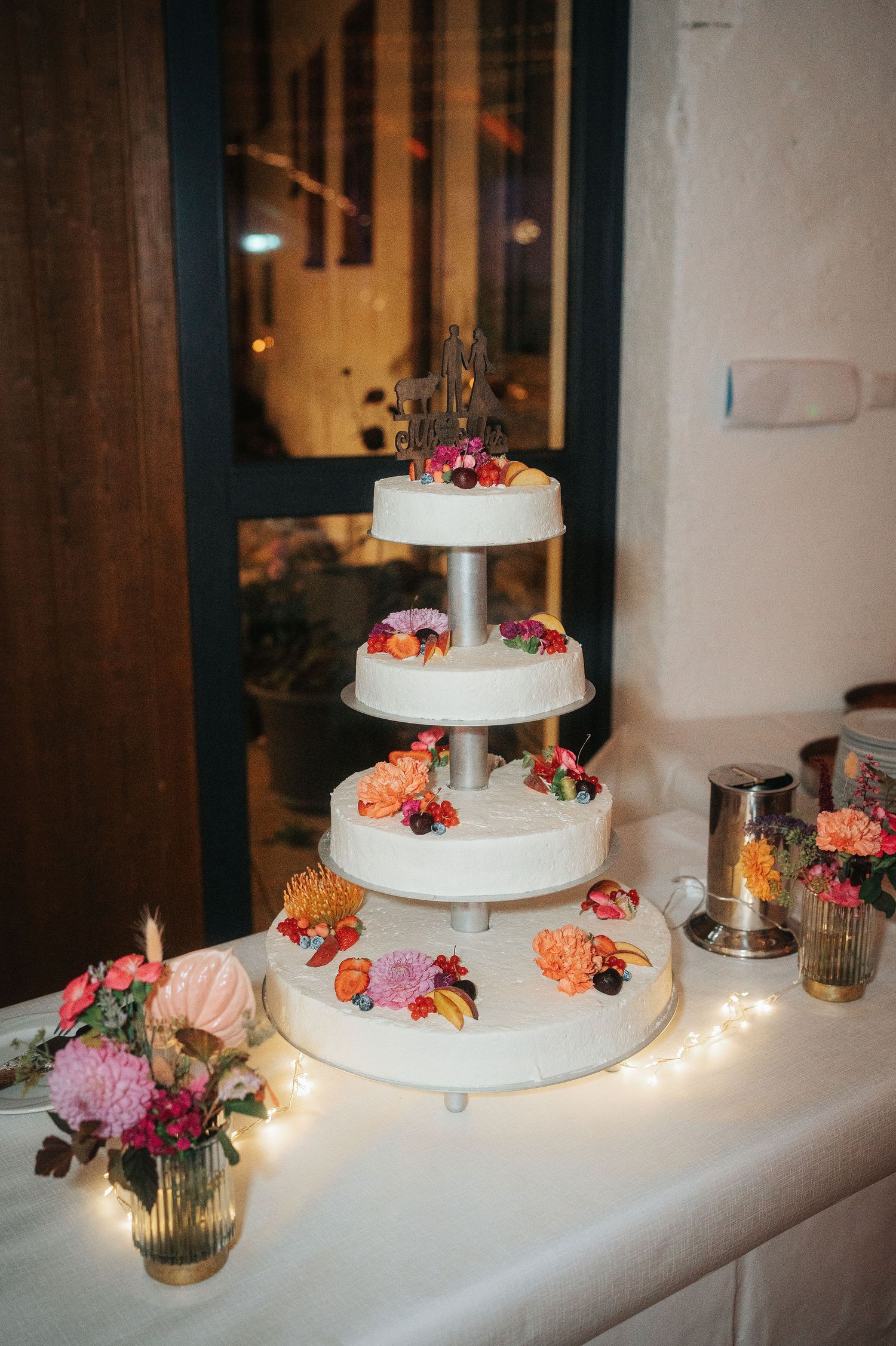 Four-tier white wedding cake decorated with colorful fresh flowers on a table with string lights, vases and a silver pitcher.