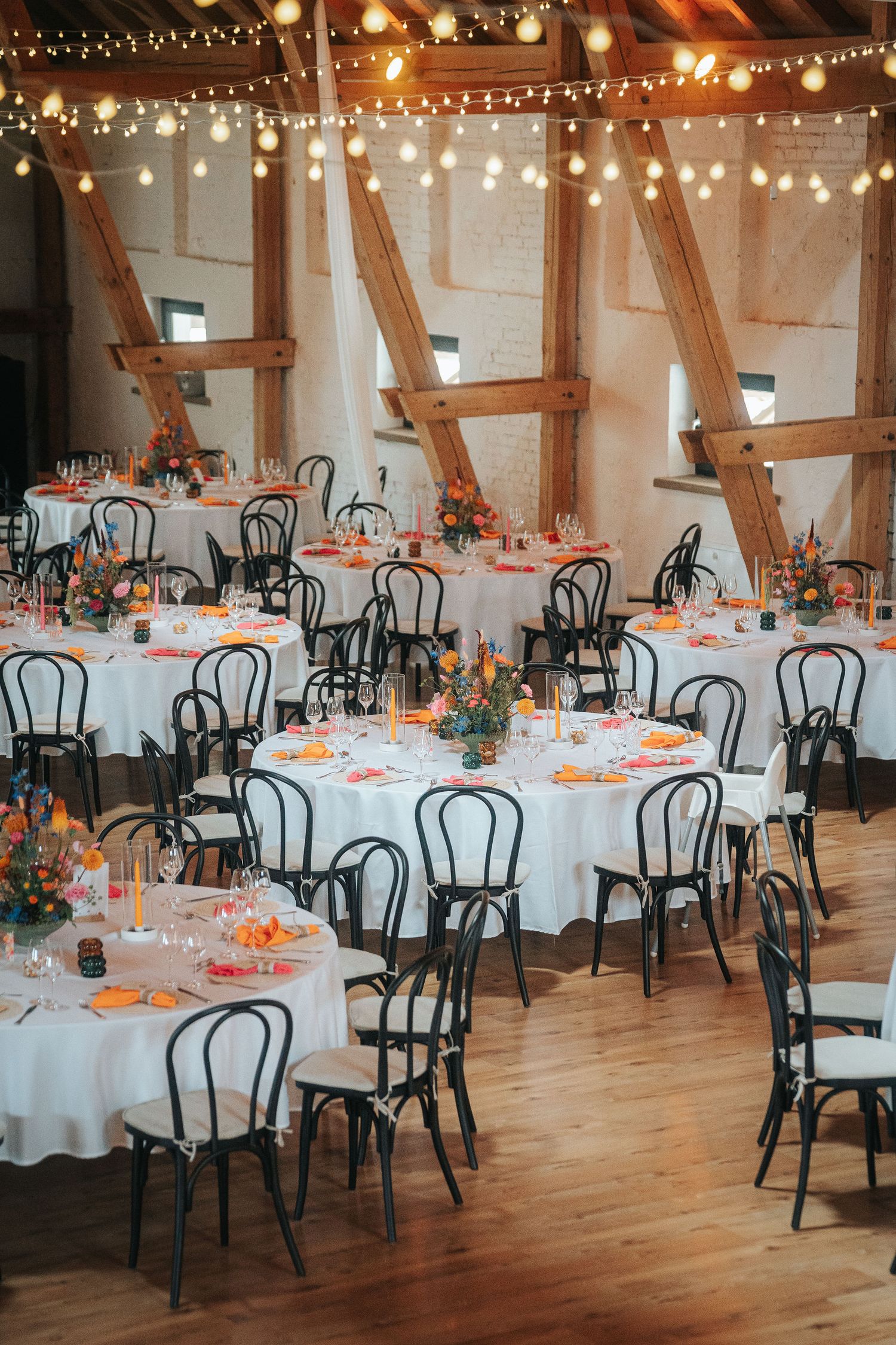 Rustic barn wedding reception with round tables, black chairs, orange floral centerpieces, string lights and exposed wooden beams.