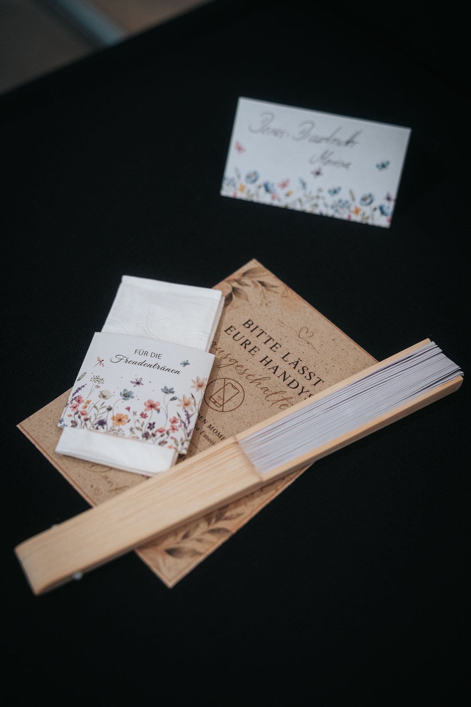 Wooden hand fan, floral tissue packet and place card on a rustic kraft card with a German phone-free request on a black table.